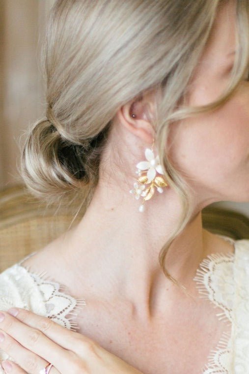 A lace clad bride with hand on heart turned to display her wedding day earrings made with enamel flowers, fresh water pearls, and gold-filled.