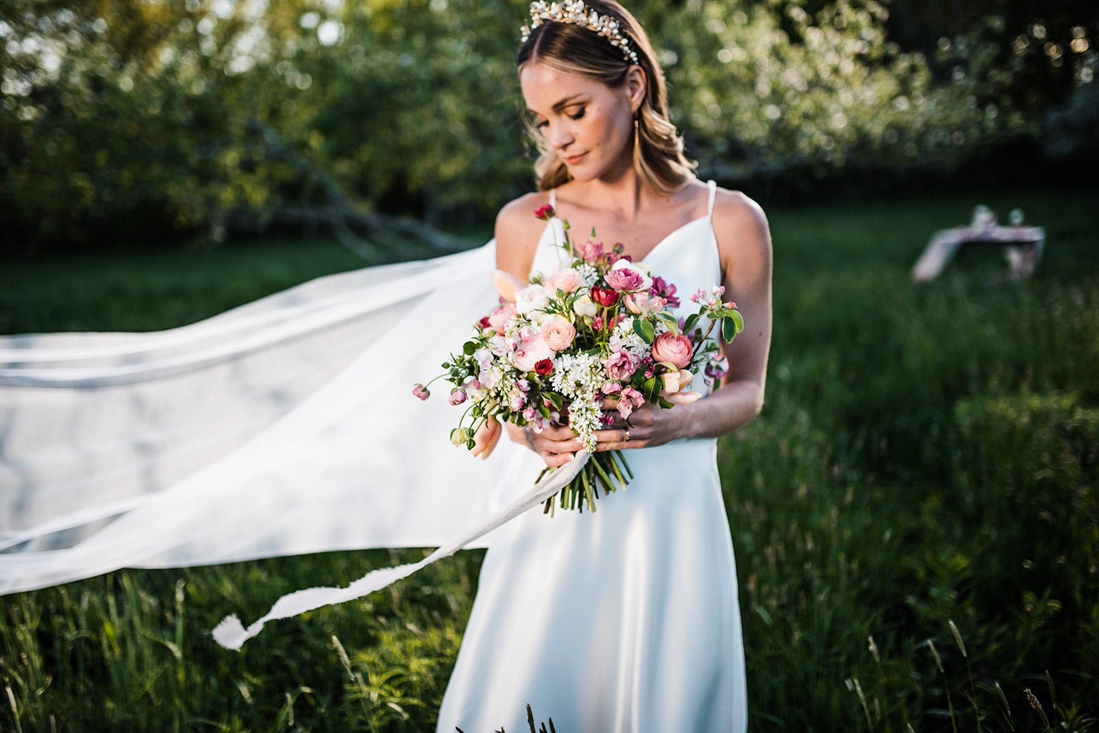 A bride with her bouquet stands in a grassy field looking down; her wedding veil flowing in the wind. 