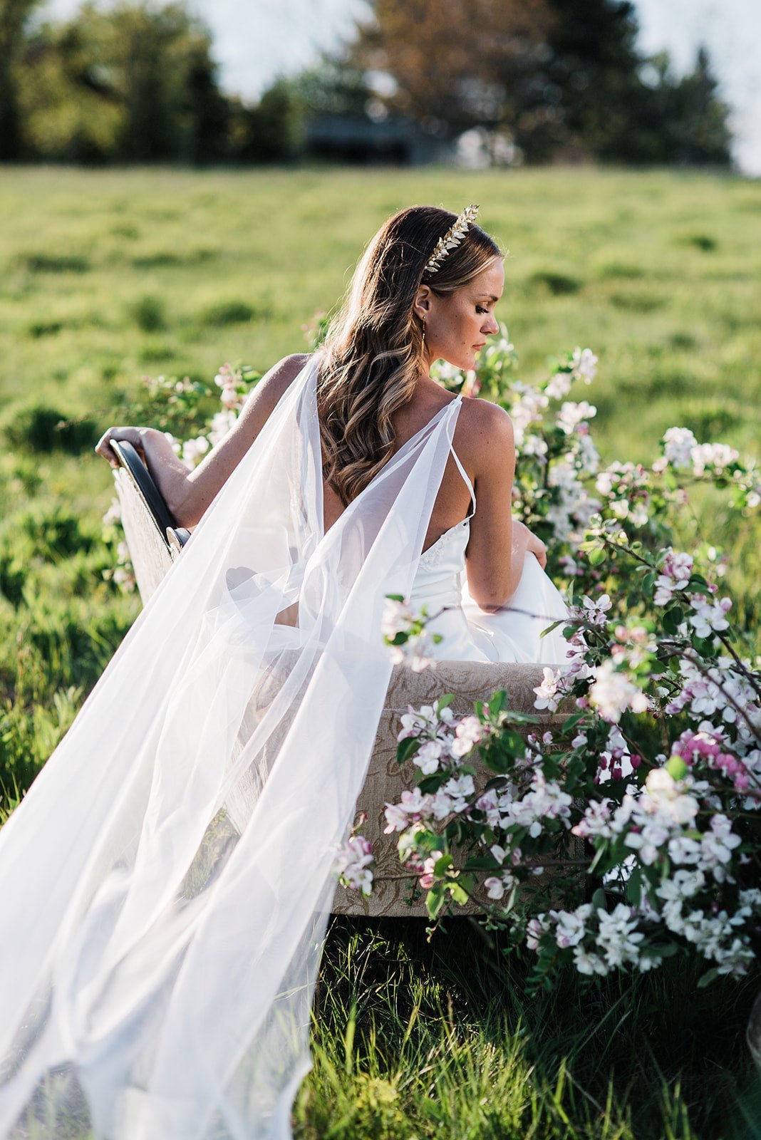 A bride sits in a field, surrounded by flowers wearing a cape-style wedding veil. 