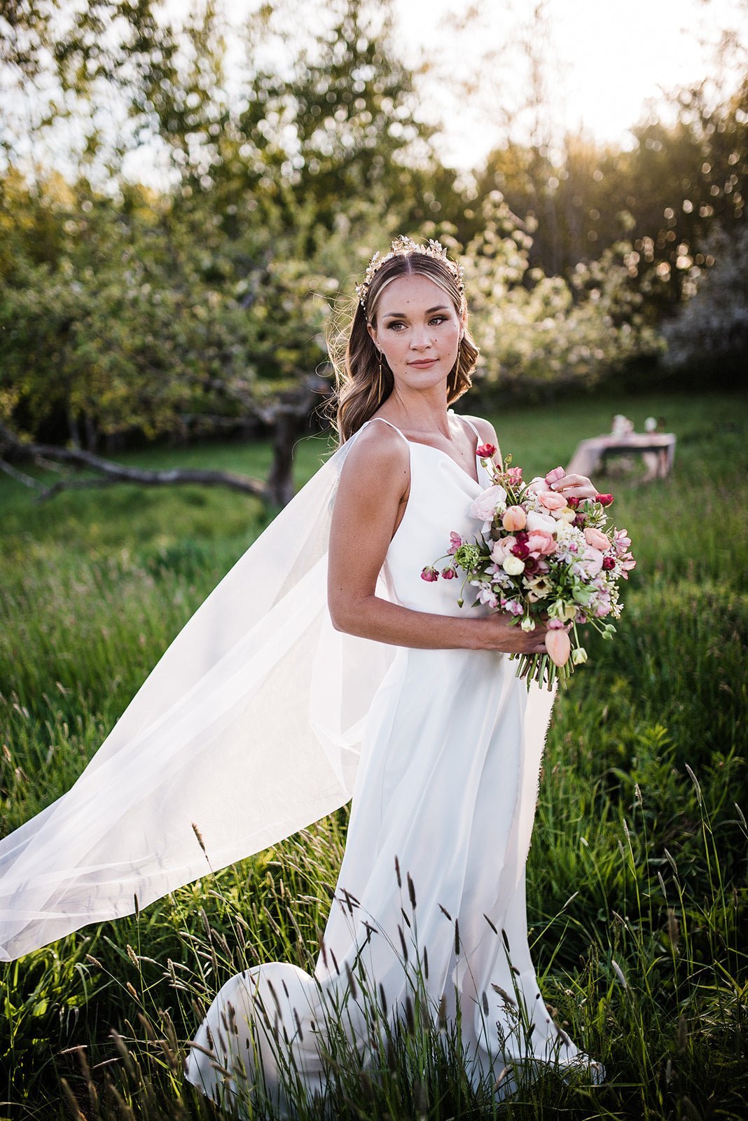 A bride stands in a field holding her bridal bouquet as her caped wedding veil flutters behind her. 