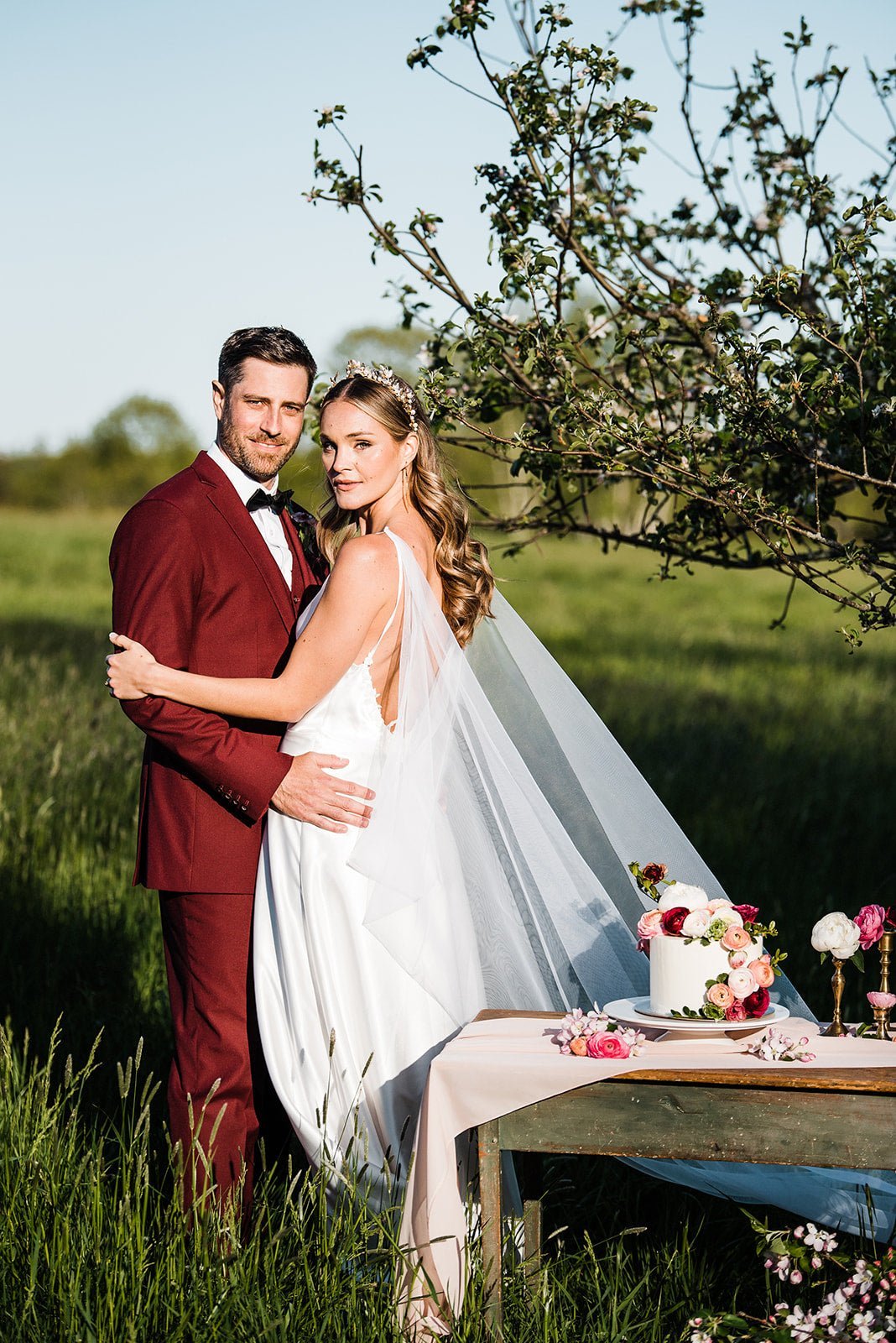 A bride and groom embrace in a field; her wedding veil is caped and flows out behind her. 