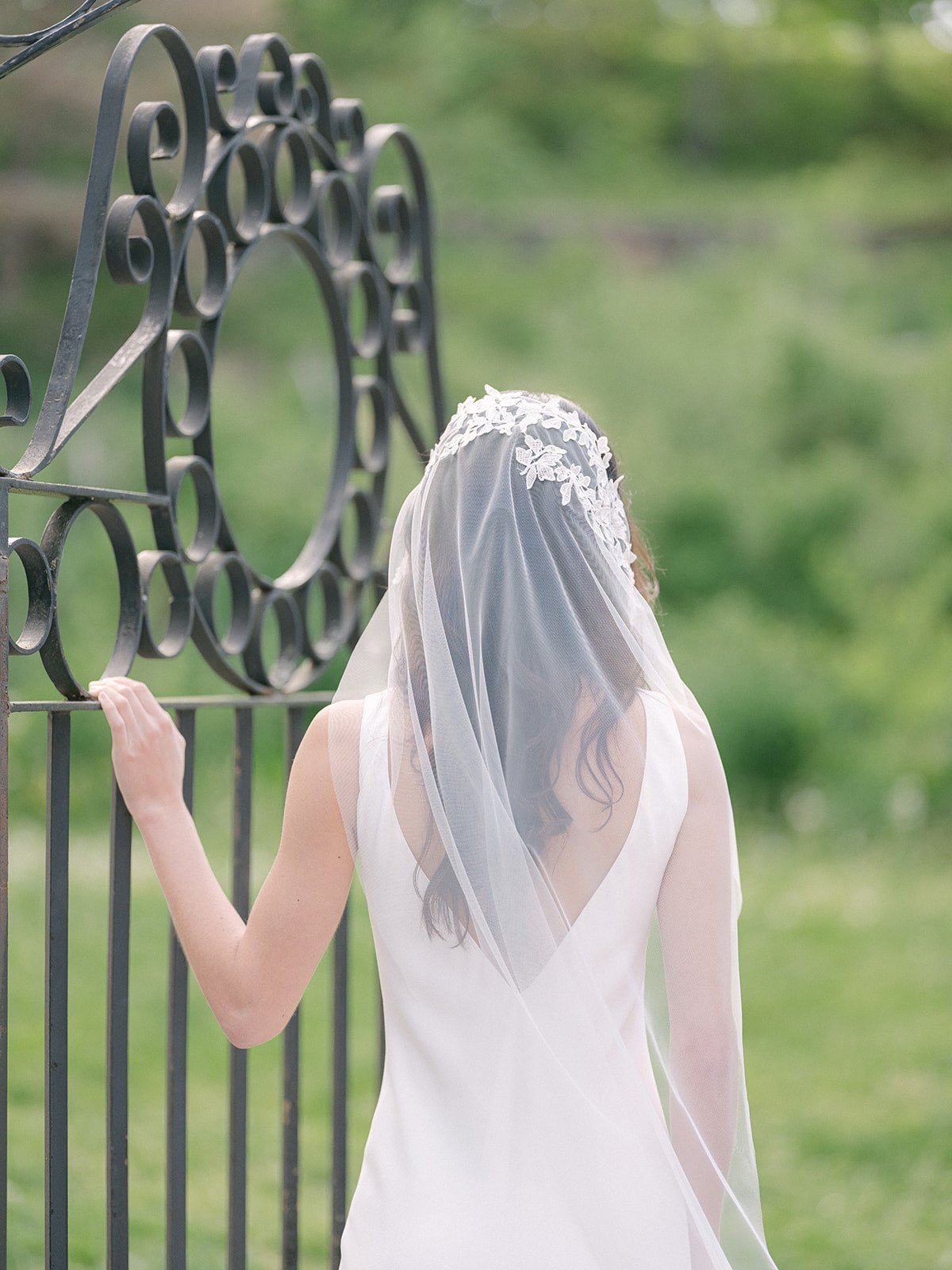 A bride holds onto a wrought iron gate as her wedding veil trails down behind her. The soft lace like a halo around the top of her head. 