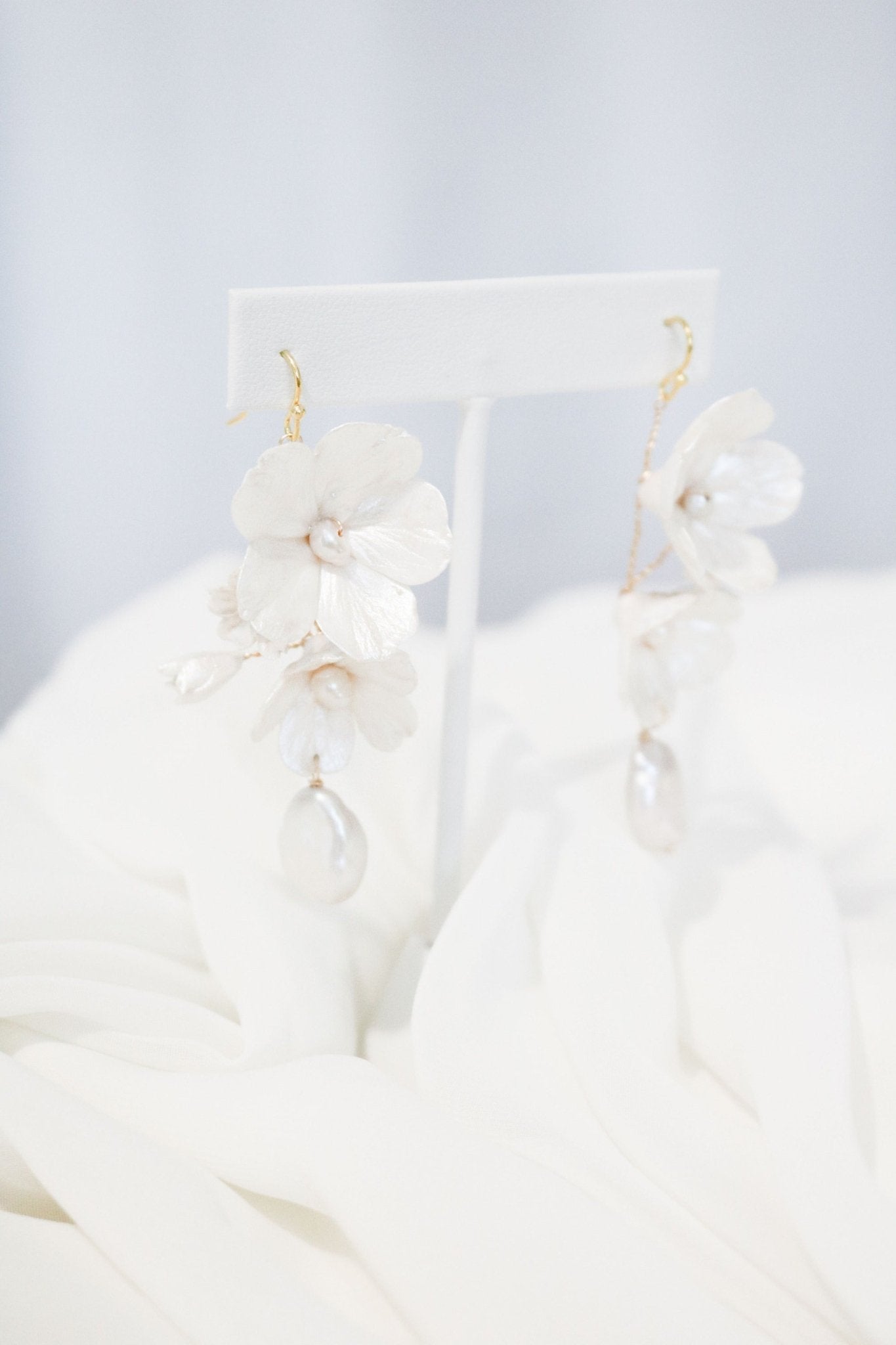 Bridal earrings in Gold with clay enamel flowers and baroque freshwater pearls hanging from a white stand over white roses. 