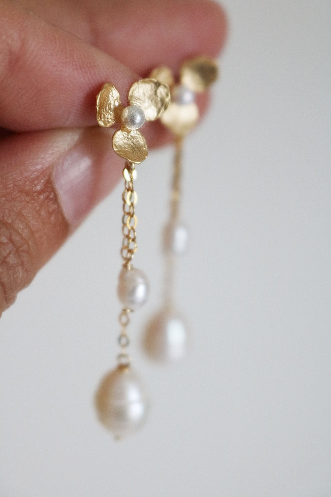 Fingers hold up a pair of elegant floral gold-filled earrings; freshwater pearls dangle down.