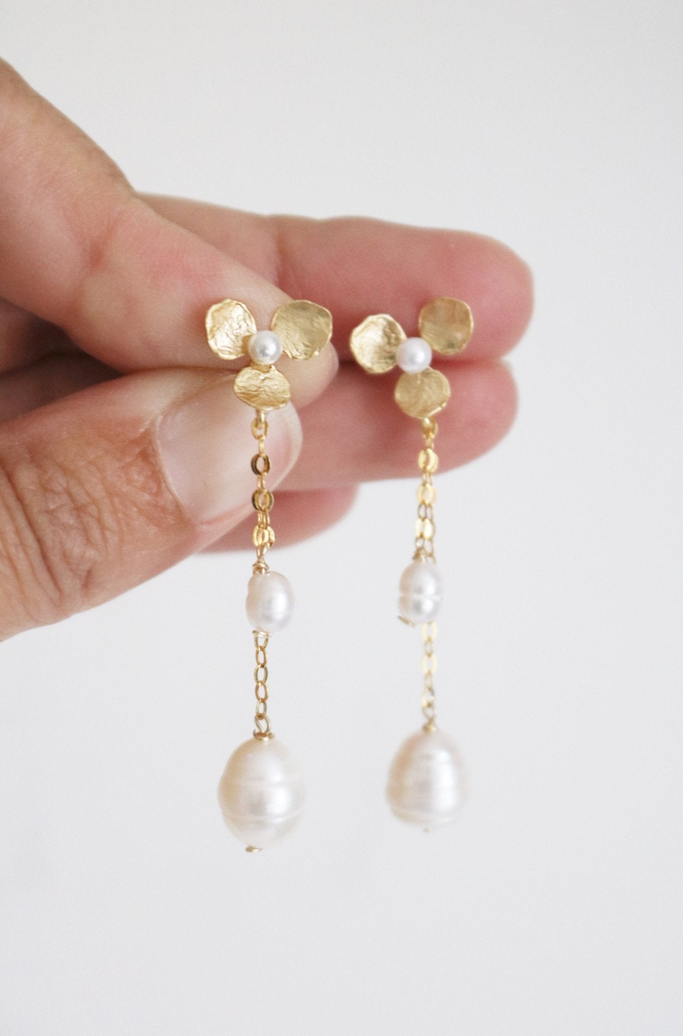 A hand showcases a pair of bridal earrings in gold-filled floral with fresh water pearls. 