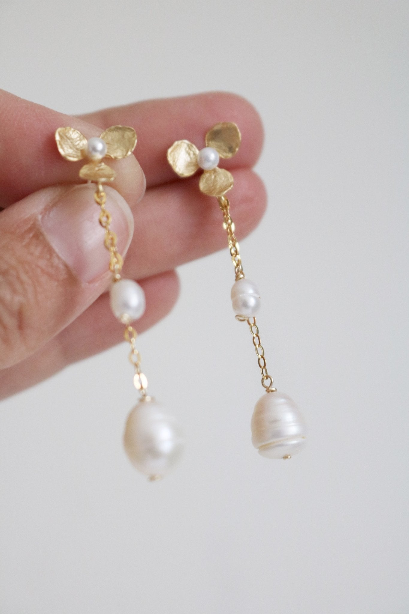 A hand showcases a pair of bridal earrings in gold-filled floral with fresh water pearls. 