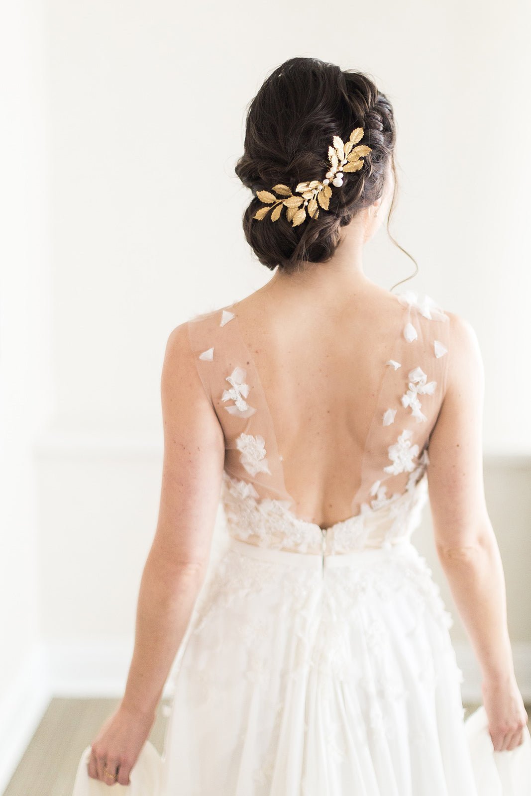 A back-turned bride in a wispy bridal gown with a braided up-do holding a golden leafy haircomb with freshwater pearls. 