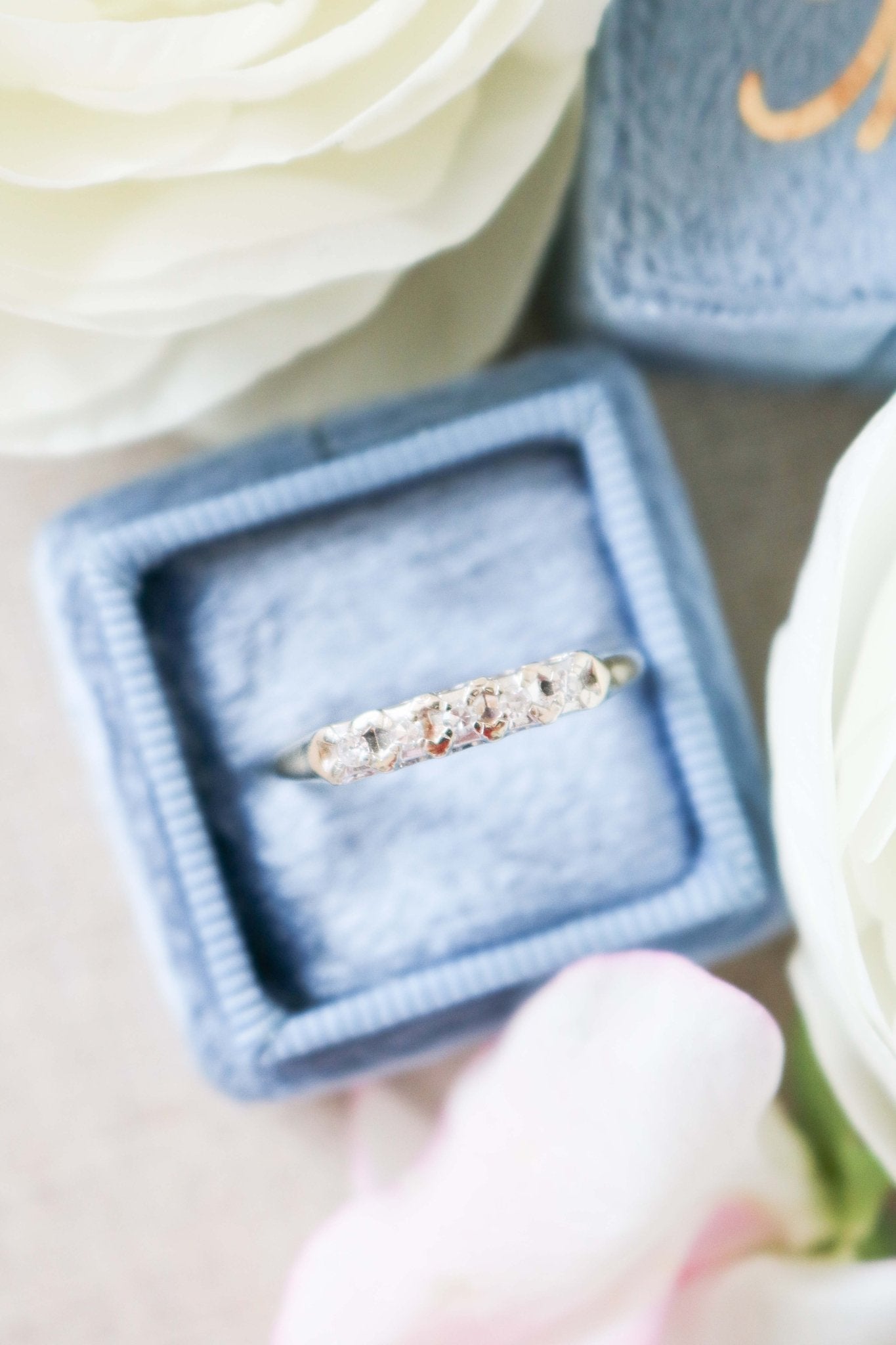 A close up of the Harmony Vintage Engagement Ring/Wedding Band in 14ct Gold with Diamonds in a ring box surrounded by white roses. 