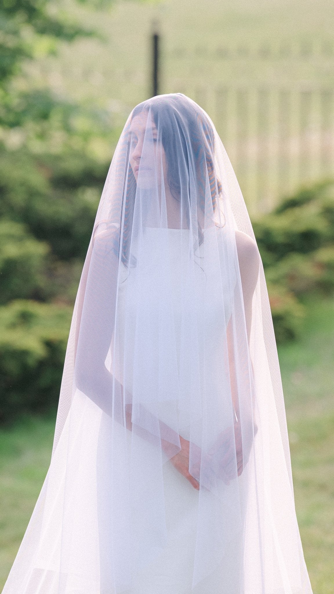 A shy bride looks poised under her full wedding veil with blusher against greenery. 