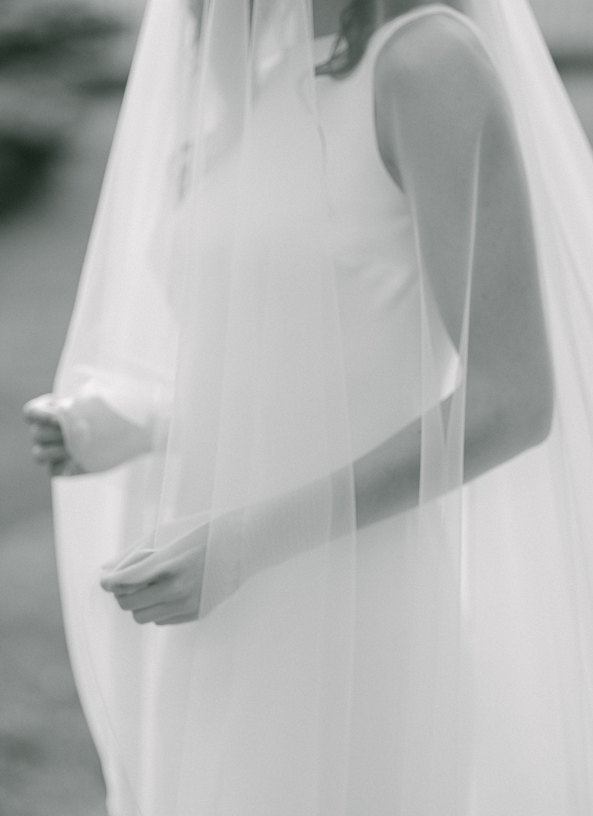 Black and white photo of torso close up of bride's hands and arms behind a long wedding veil. 