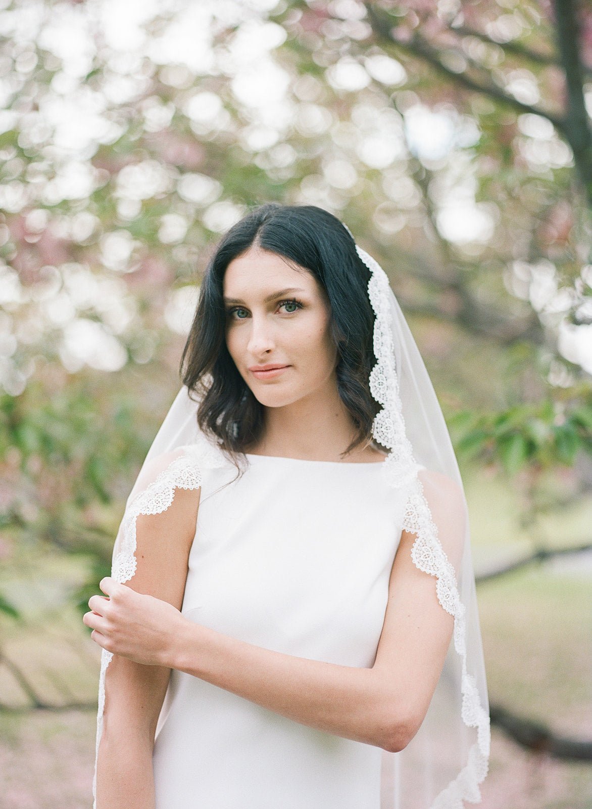 A bride in nature dons a white lace, scallop-edged veil against dark hair.