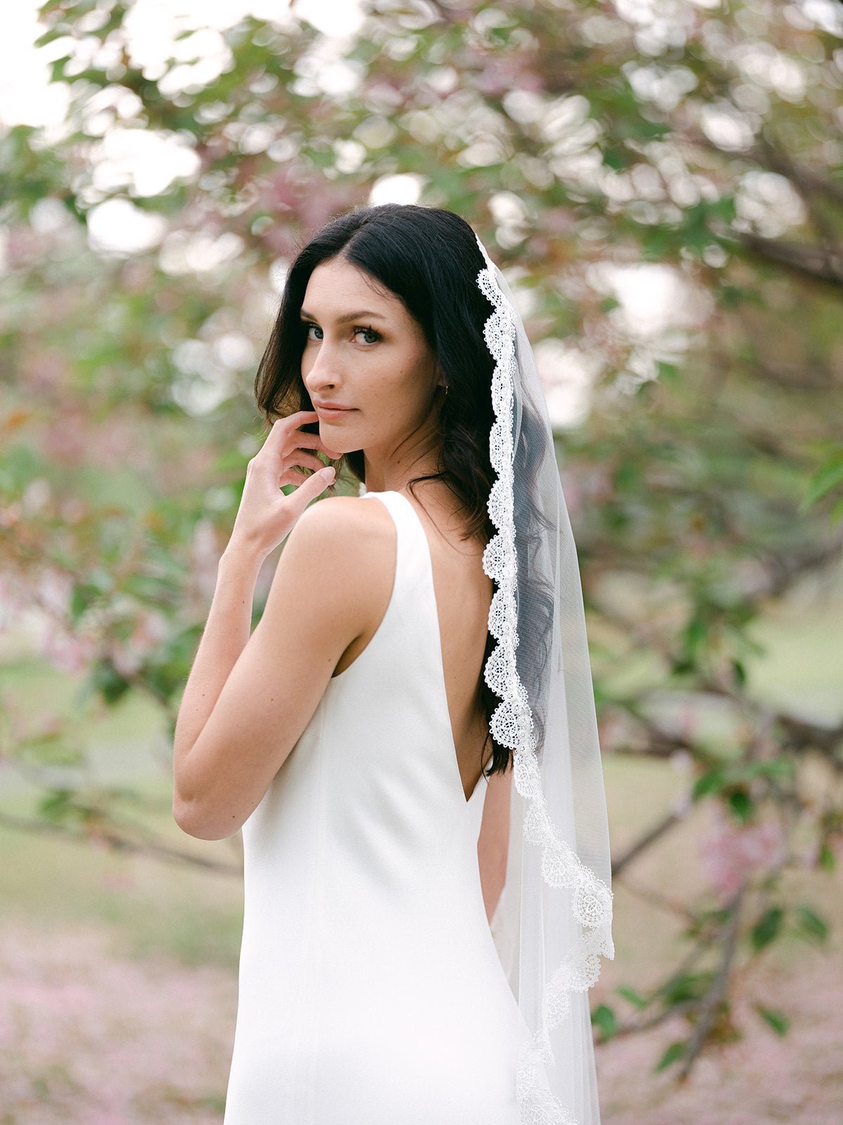 A bride in white looking over her shoulder wearing a wedding dress with a sheer lace veil.