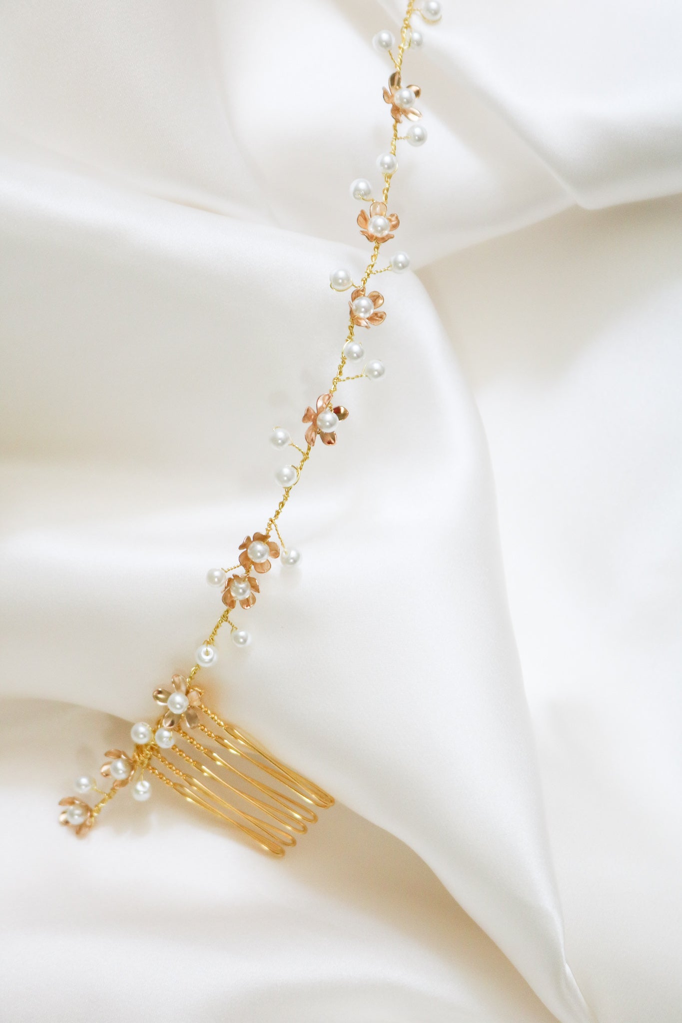 A golden bridal hairvine with flowers and freshwater pearls lays on a bed of white satin. 