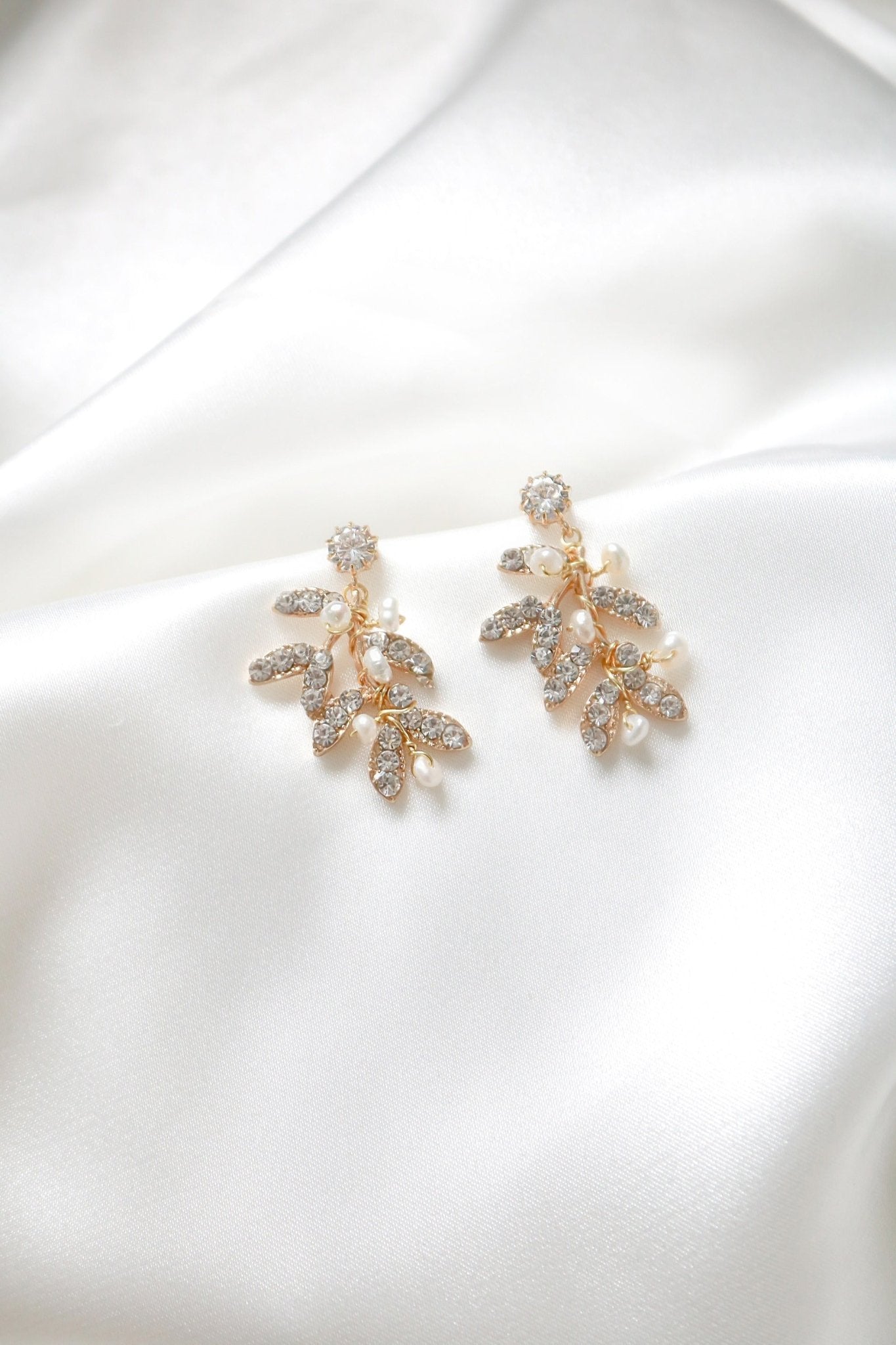 A pair of gold-filled leafy studs with crystals and freshwater pearls sitting on a background of white satin. 