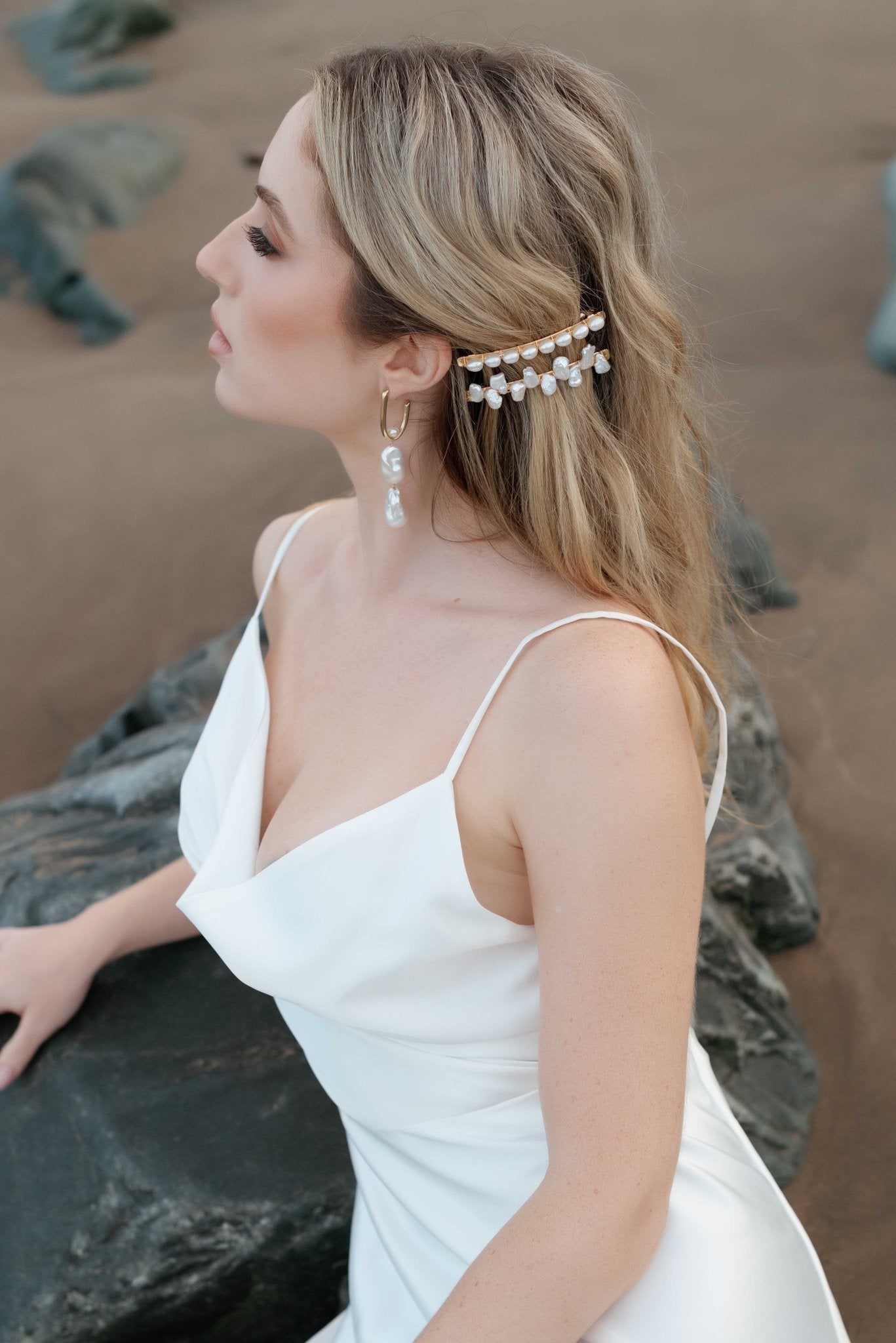 Elegant bride lounges against a rock at the beach keeps hair aside with gold and pearl wedding barrettes. She also displays a double baroque pearl hanging earring. 