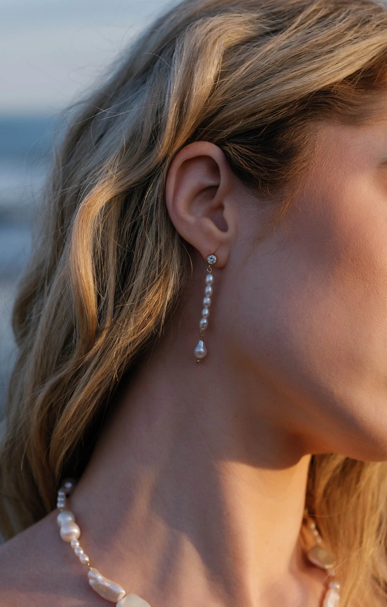 A close up of a gold-filled freshwater pearl drop bridal earring with crystal stud dangling from a bride's ear with ocean background. Organic pearl jewelry & Bridal earrings handmade by BIPOC bridal designer