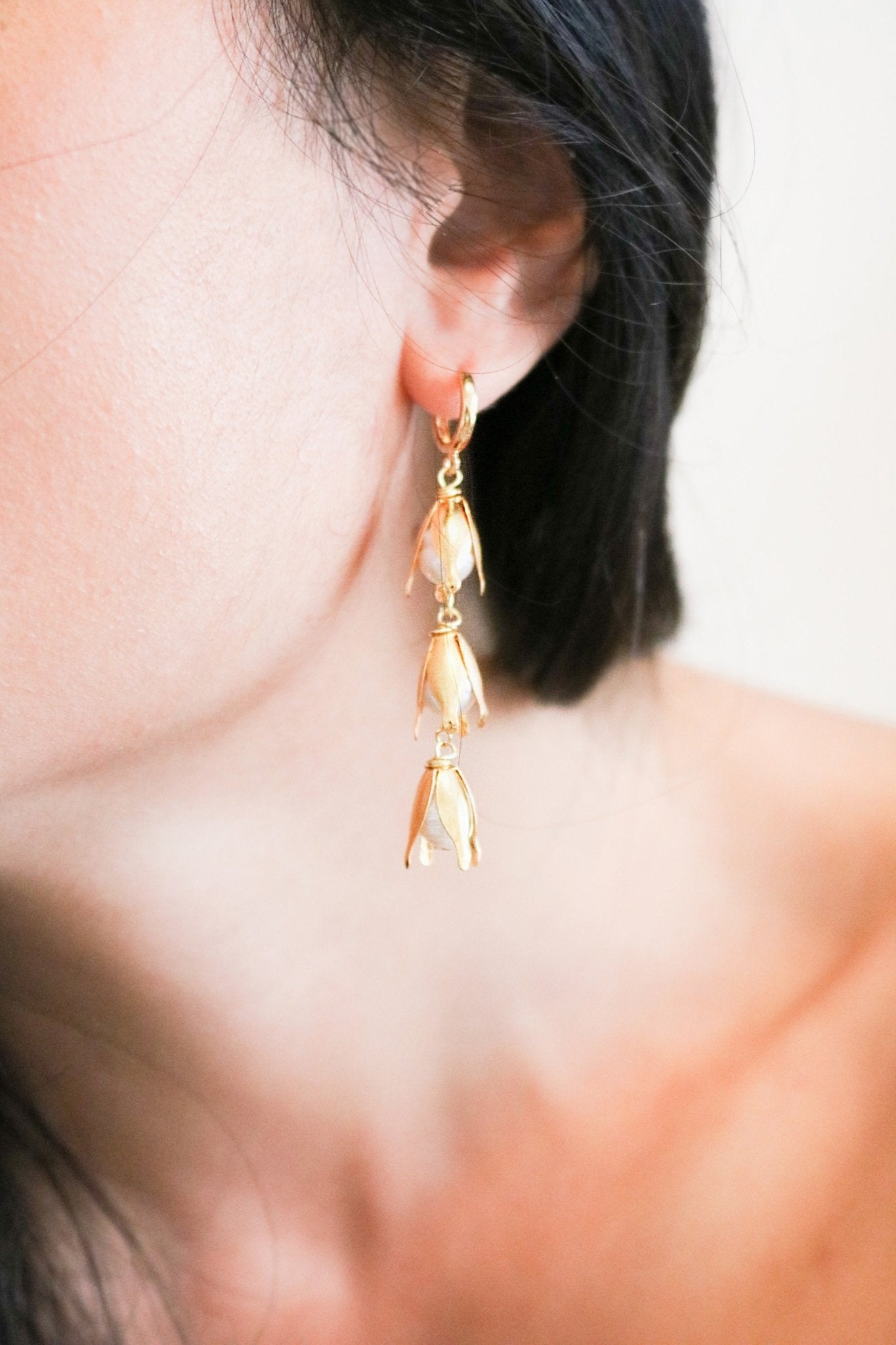 A gold-filled flower bud dangle earring with freshwater pearls on a huggie-style hoop dangles from an earlobe. 