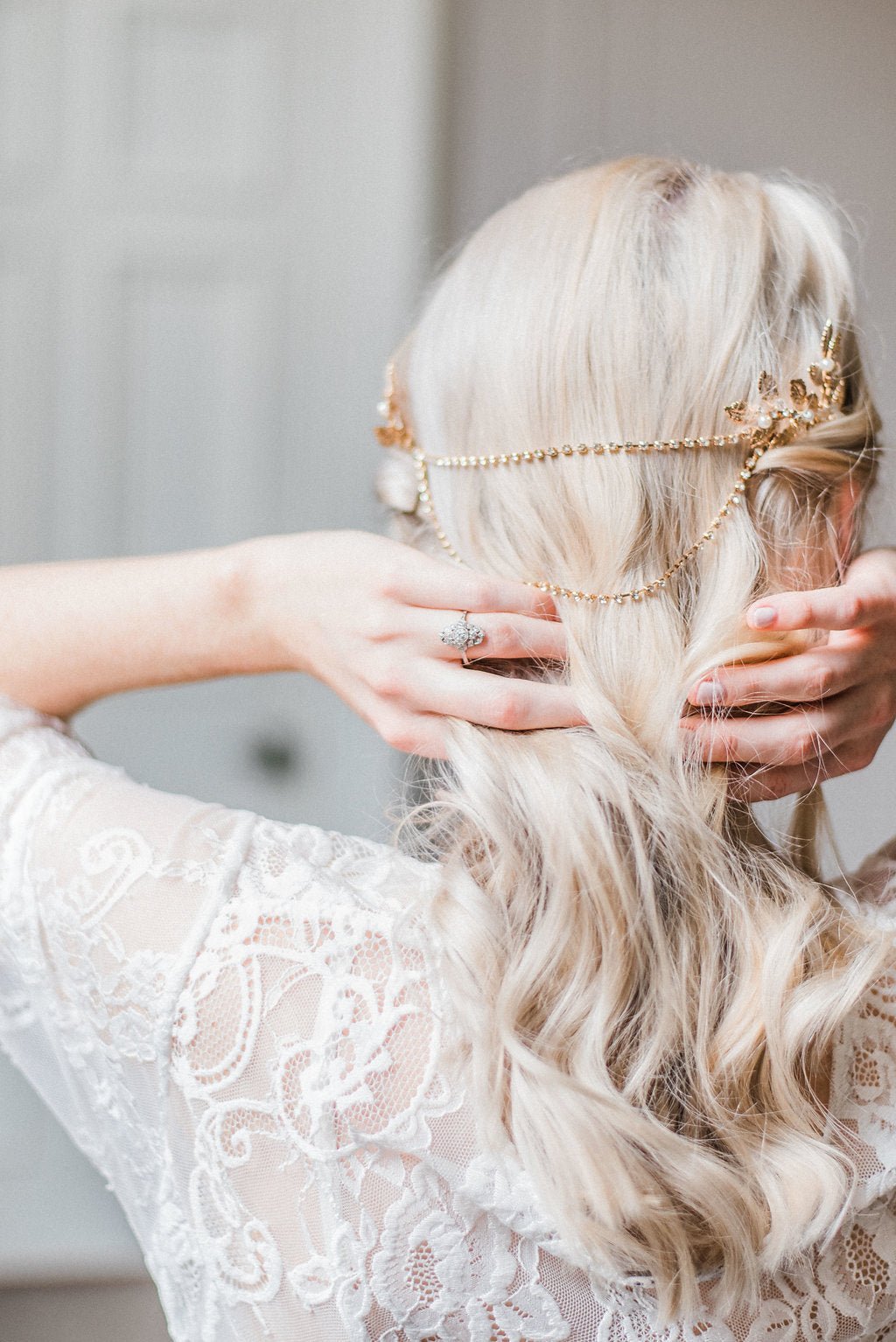 The back of a bride with white lacey wedding dress holds back of her neck while wearing a gold leafed pearl bridal hair comb with connecting crystal chains on blurred background. Bridal hair accessories & handmade Canadian accessories by BIPOC bridal jewelry designer
