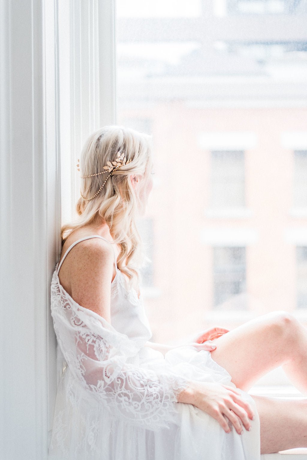 A modern bride sits on a windowsill looking out. Her lacey shawl and white wedding dress bringing out the shine of crystal from her golden bridal hair accessory. 