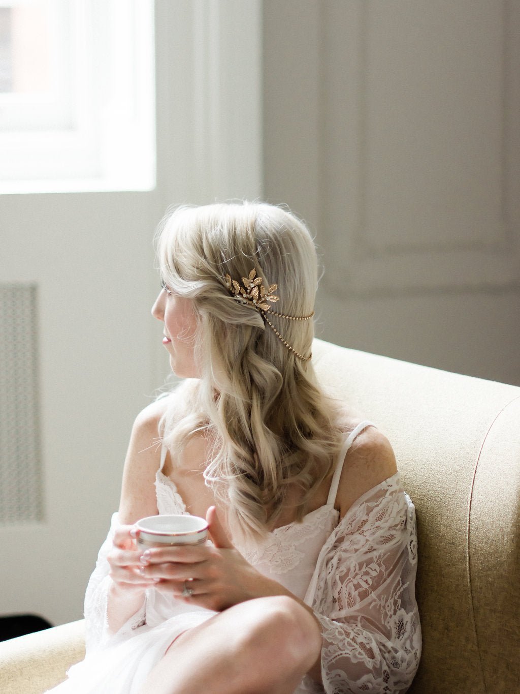 A modern bride casually sits with a cup of tea wearing a white lace shawl, white wedding gown, and a gold and crystal bridal hairpiece with freshwater pearls. 