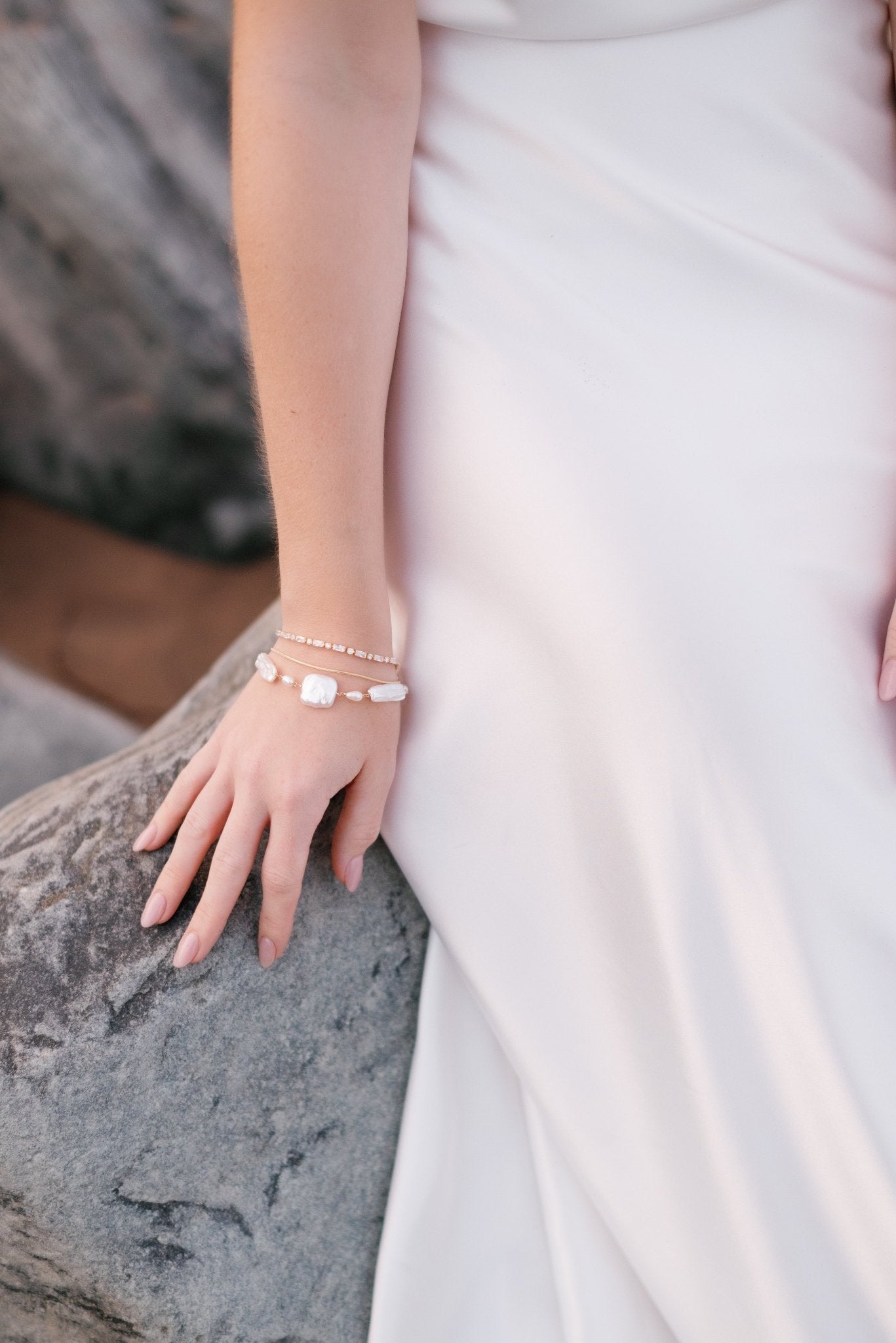 A bride sits on a beach rock in a sleek wedding gown. She wears three bracelets stacked for heightened allure; gold filled crystal, simple strand, and thick freshwater pearl.