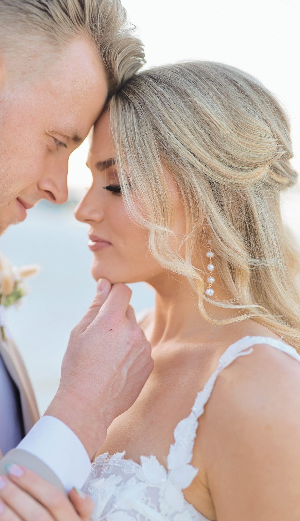 A groom hold's his bride's chin; her gold freshwater pearl dangle earrings peeking through her blonde curls. 