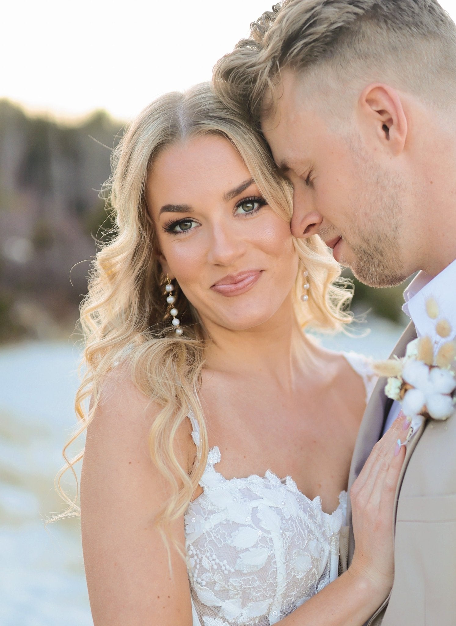 A bride and groom stand on a beach together; she displays a pair of long statement gold and freshwater pearl dangle earrings. 