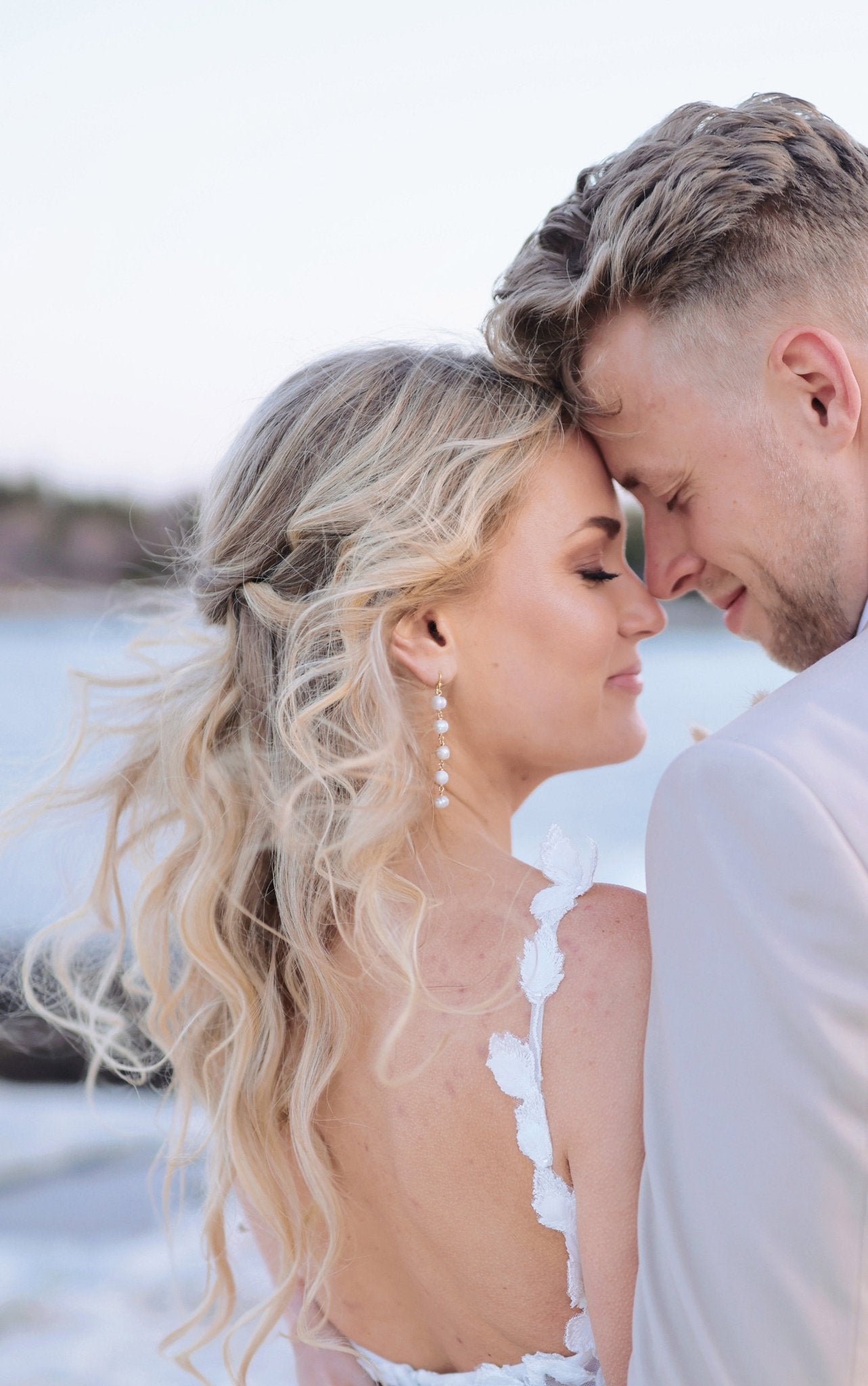 A bride and groom touch foreheads by the ocean; she is wearing a gold dangle statement freshwater pearl bridal earring. 