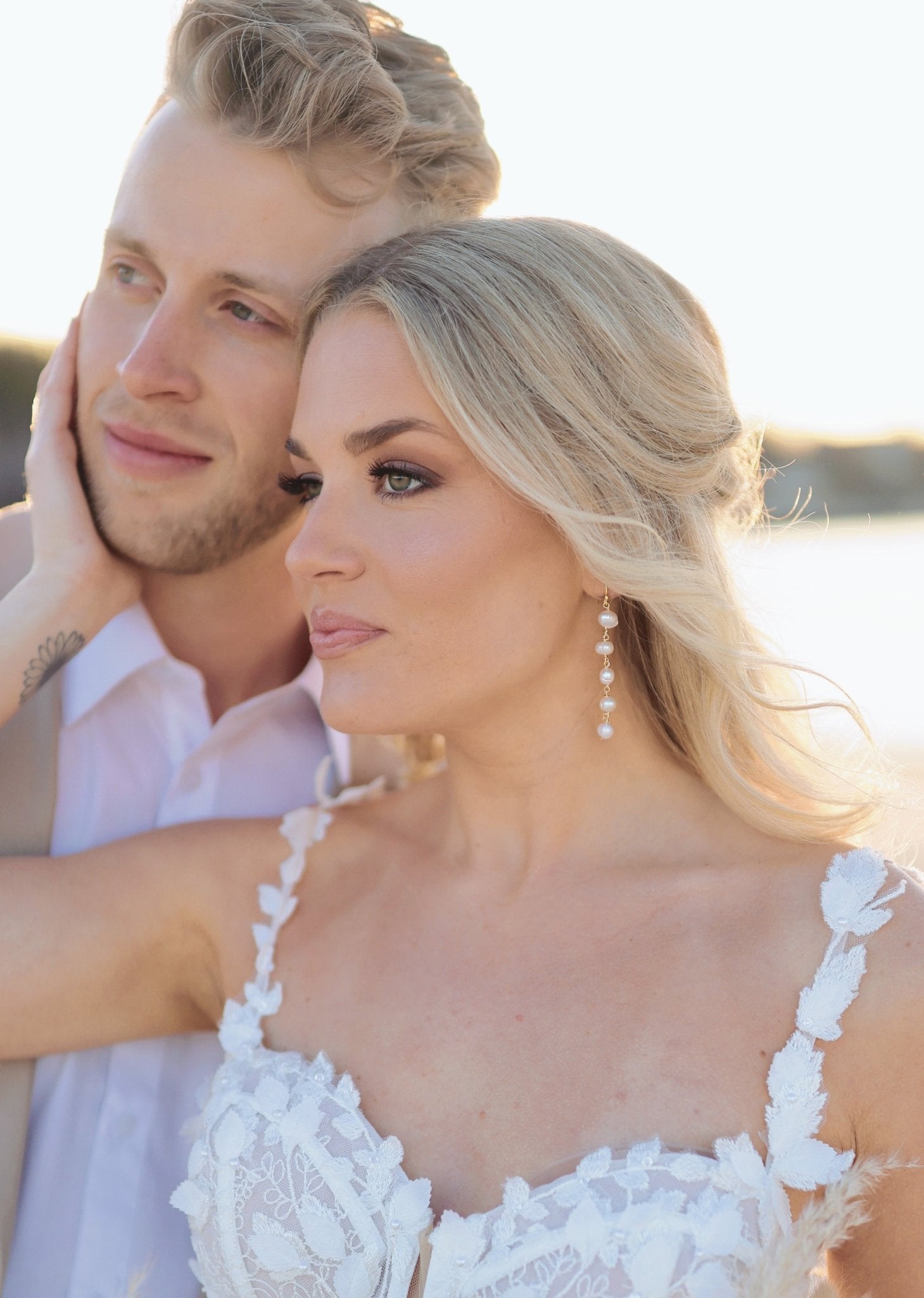 A bride cups her grooms face in her hand as they look off to the side; she has a lacey bridal gown and a gold dangle, freshwater pearl statement bridal earring. 