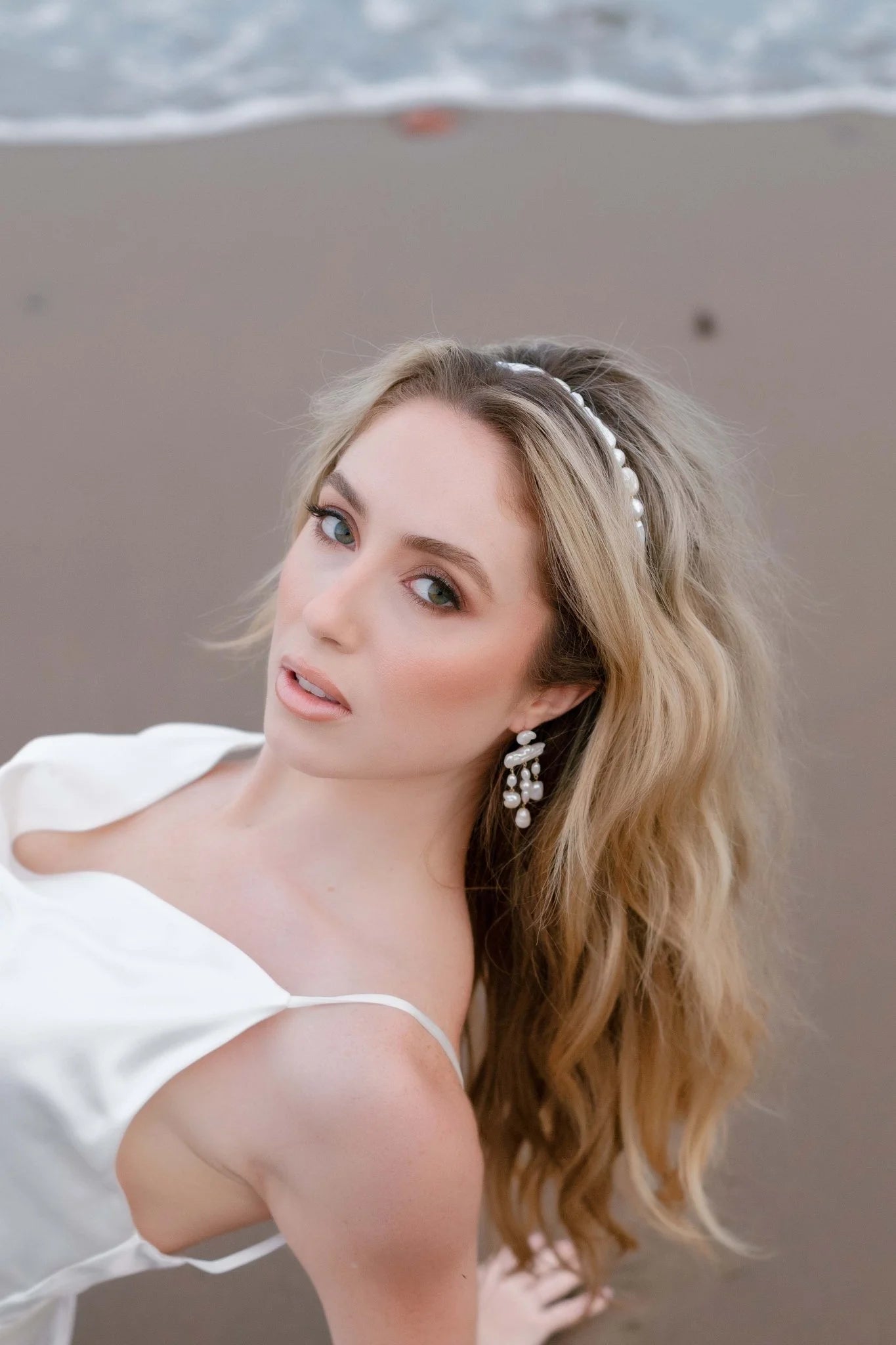 Bride leaning back on beach sand in a white wedding gown accessorized with freshwater pearl bridal headband and earring set. Organic pearl accessories, Pearl Bridal Hair Accessories, & handmade bridal jewelry Canada