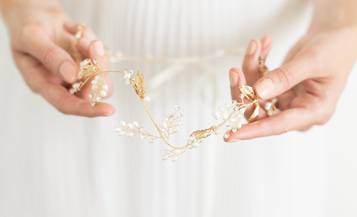 A golden bridal hairvine with crystals and freshwater pearls is held against a white wedding dress.