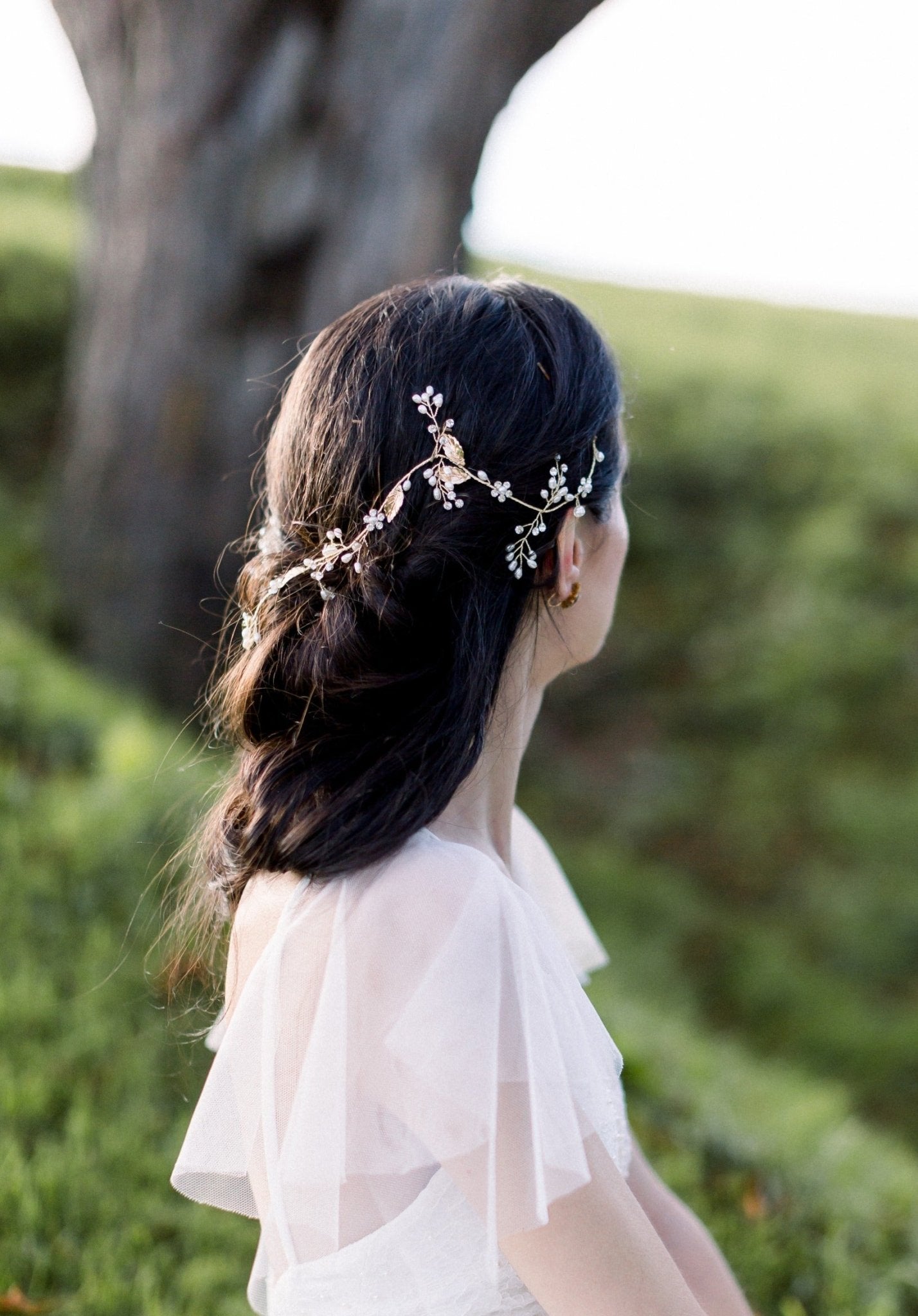 A bride looks out of frame at the top of a hill to showcase her partial up-do graced with a golden bridal hairvine with freshwater pearls. 