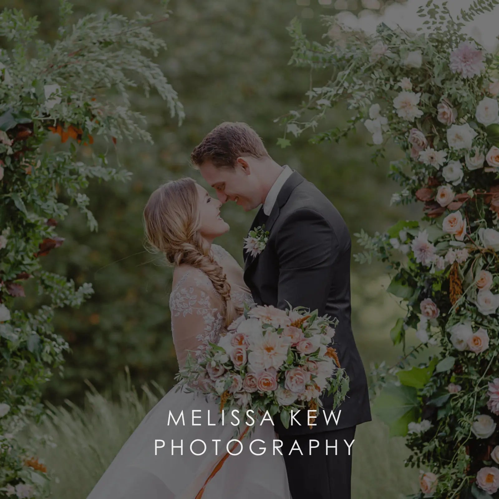 A bride and groom touch noses surrounded by roses and greenery while holding a large spring wedding flower bouquet.
