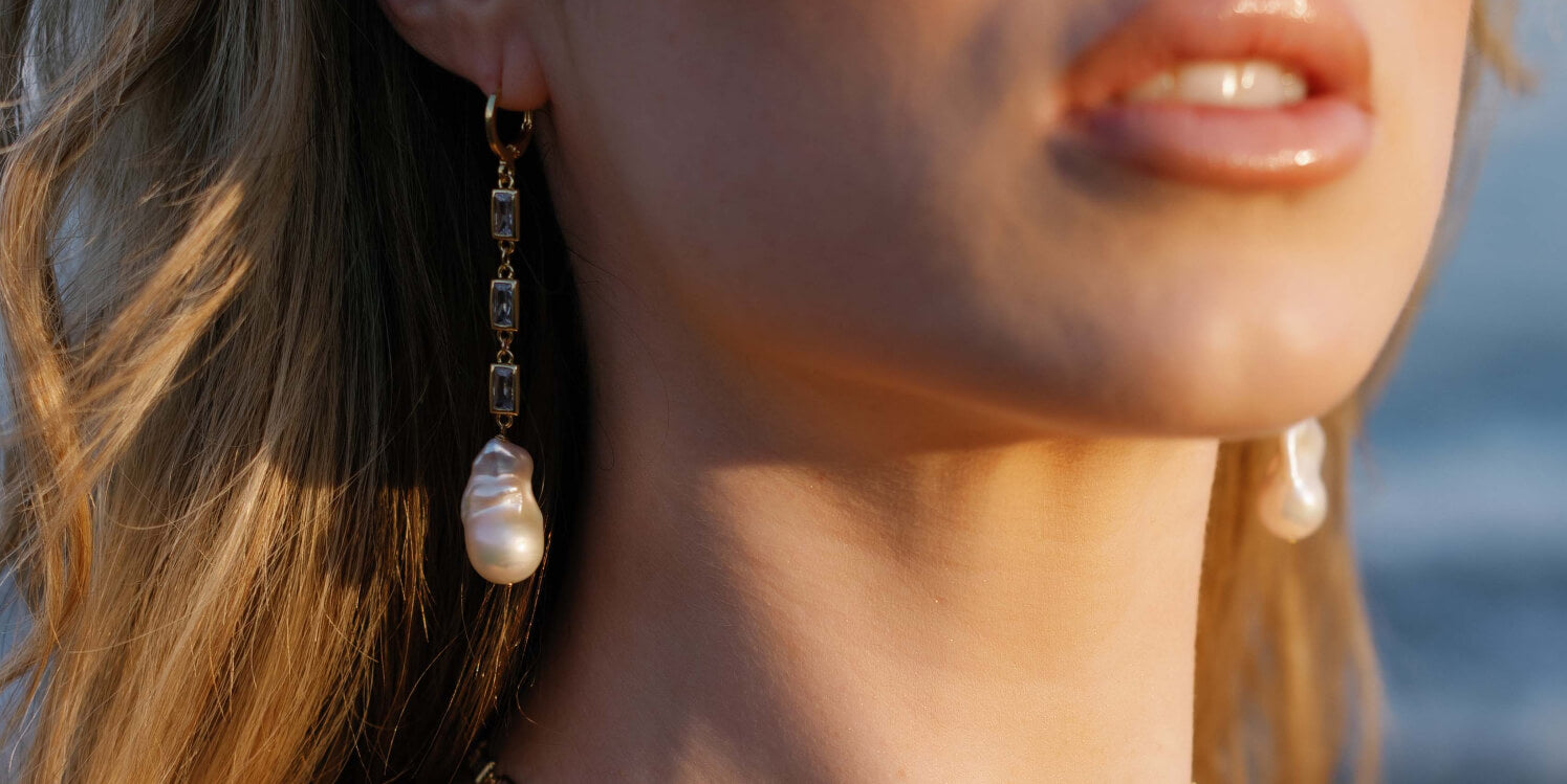 Close up of a bride wearing a gold-filled crystal baroque freshwater pearl dangle earring with ocean background. Handmade bridal jewelry, pearl jewelry sets, & statement bridal earrings by BIPOC wedding designer