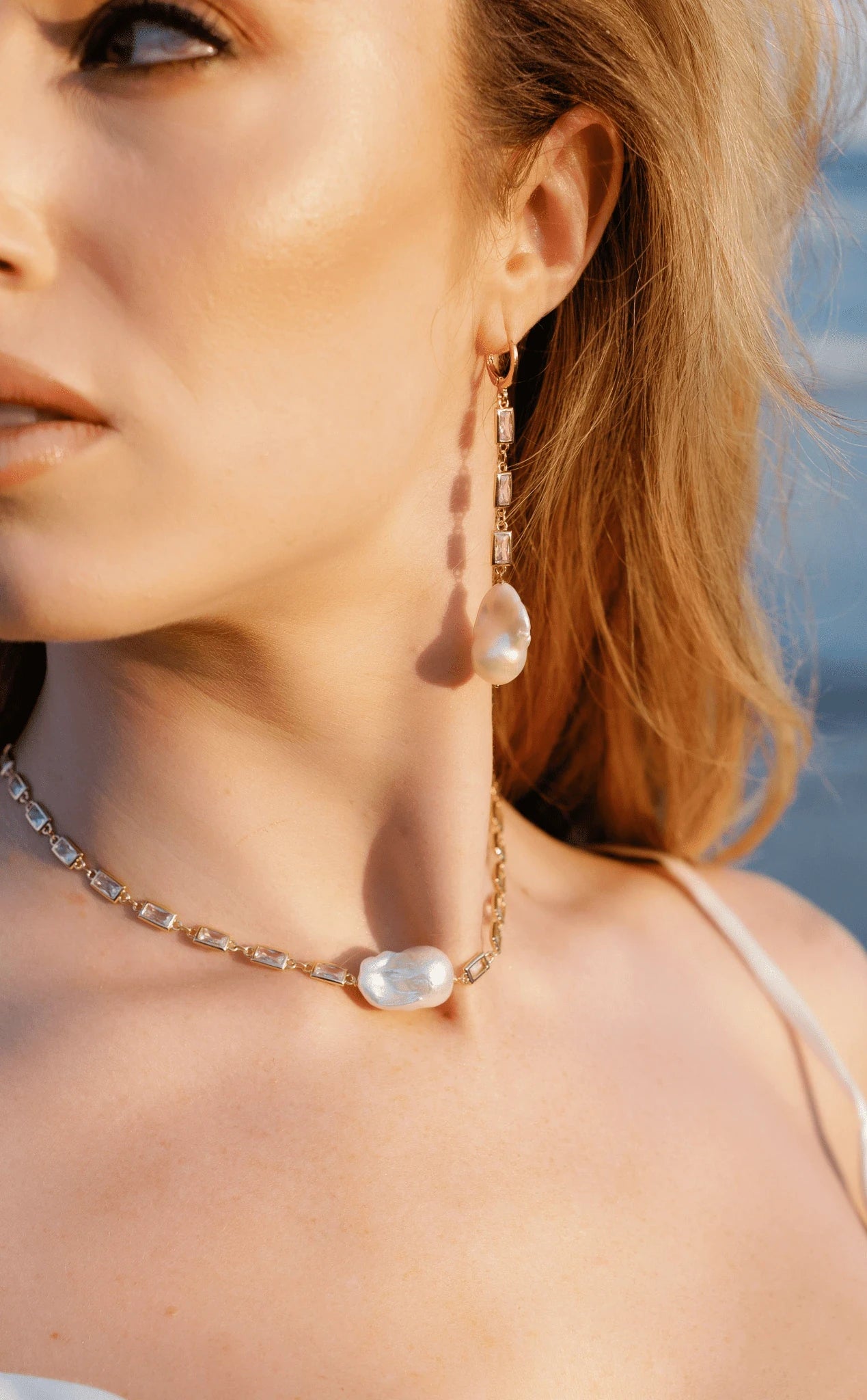 A bride wearing a gold-filled crystal baroque freshwater pearl dangle earring and matching necklace with ocean background. Affordable bridal jewelry, pearl jewelry sets, statement bridal earrings by BIPOC wedding designer