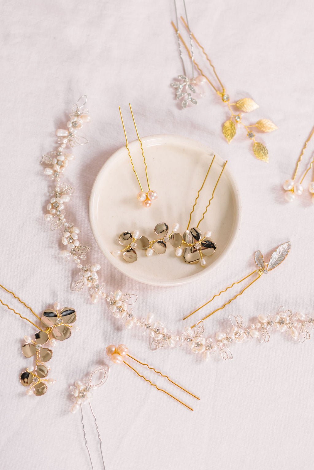 A gold crystal and freshwater pearl bridal hairvine surrounded by hairpins. Some sit in a jewelry dish.