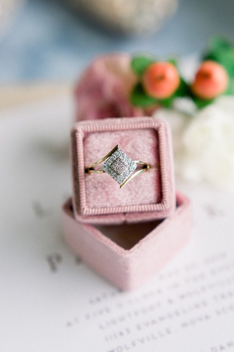 A close up of a Vintage Engagement Ring in 10k Yellow & White Gold with Diamonds sits in a pink square ring box on top of a wedding invitation. 