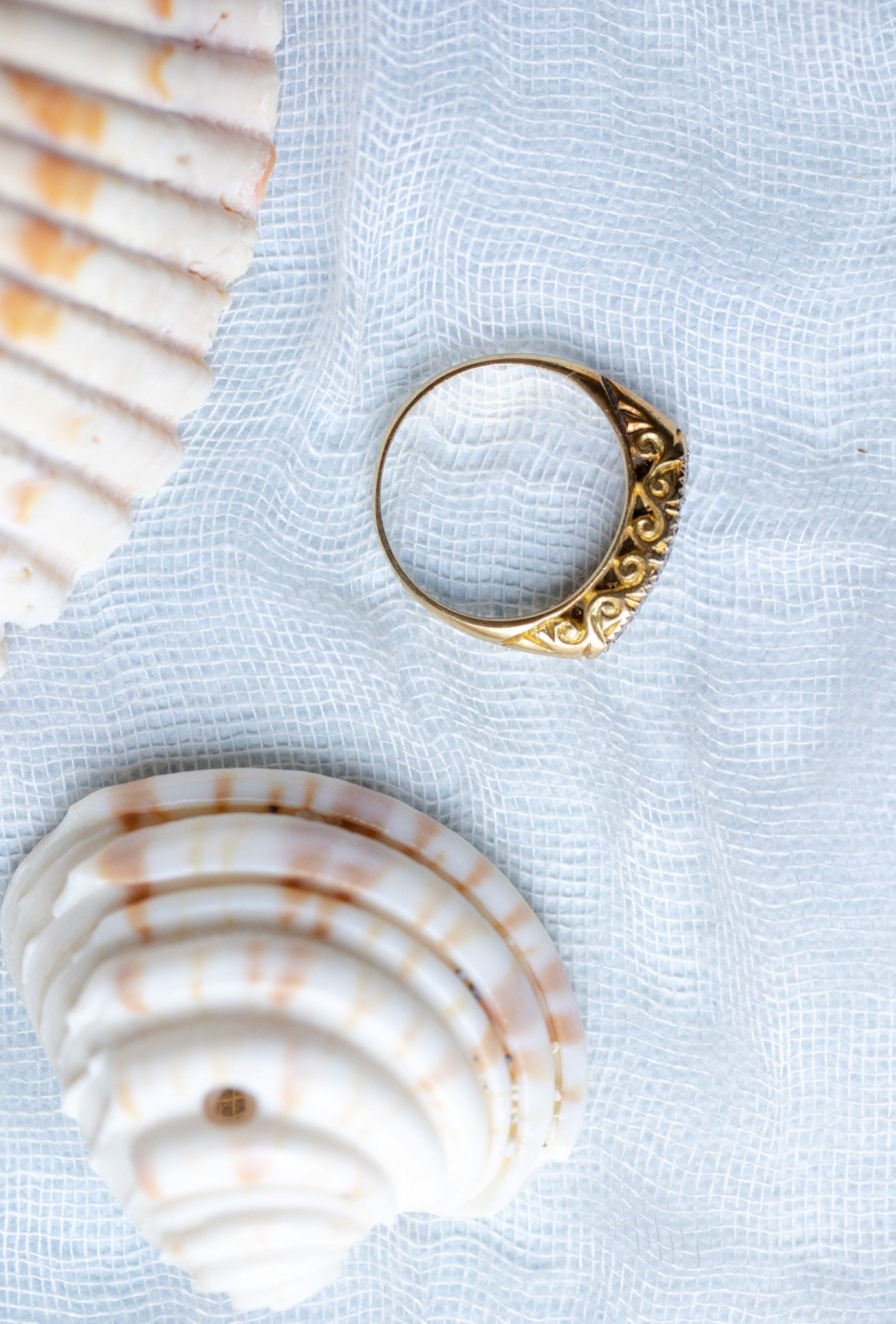 A close up of the side view of a 18k gold diamond vintage engagement ring with seashells on a pale blue fabric. 