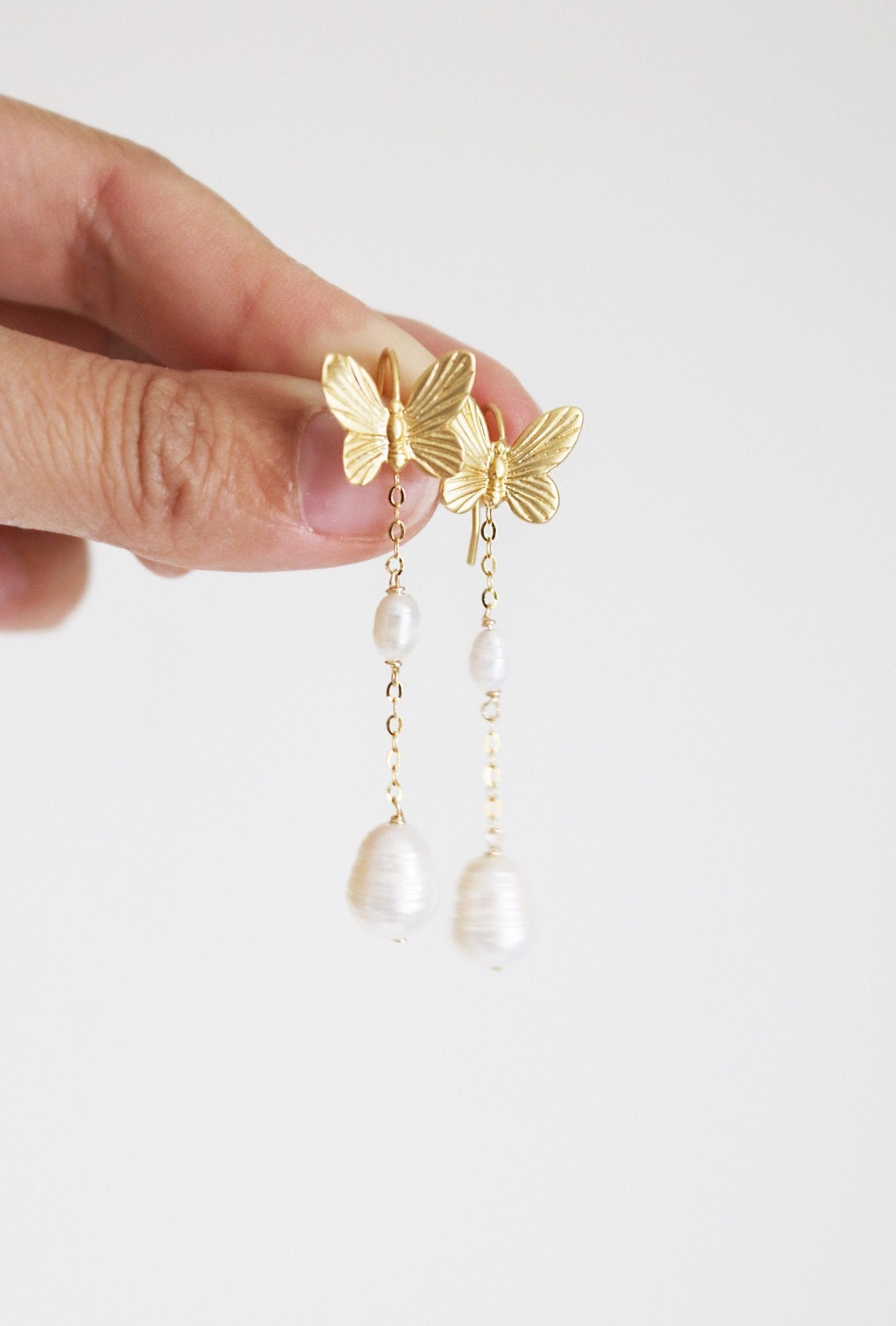 A pair of butterfly gold-filled earrings with freshwater pearls being held be two fingers. 