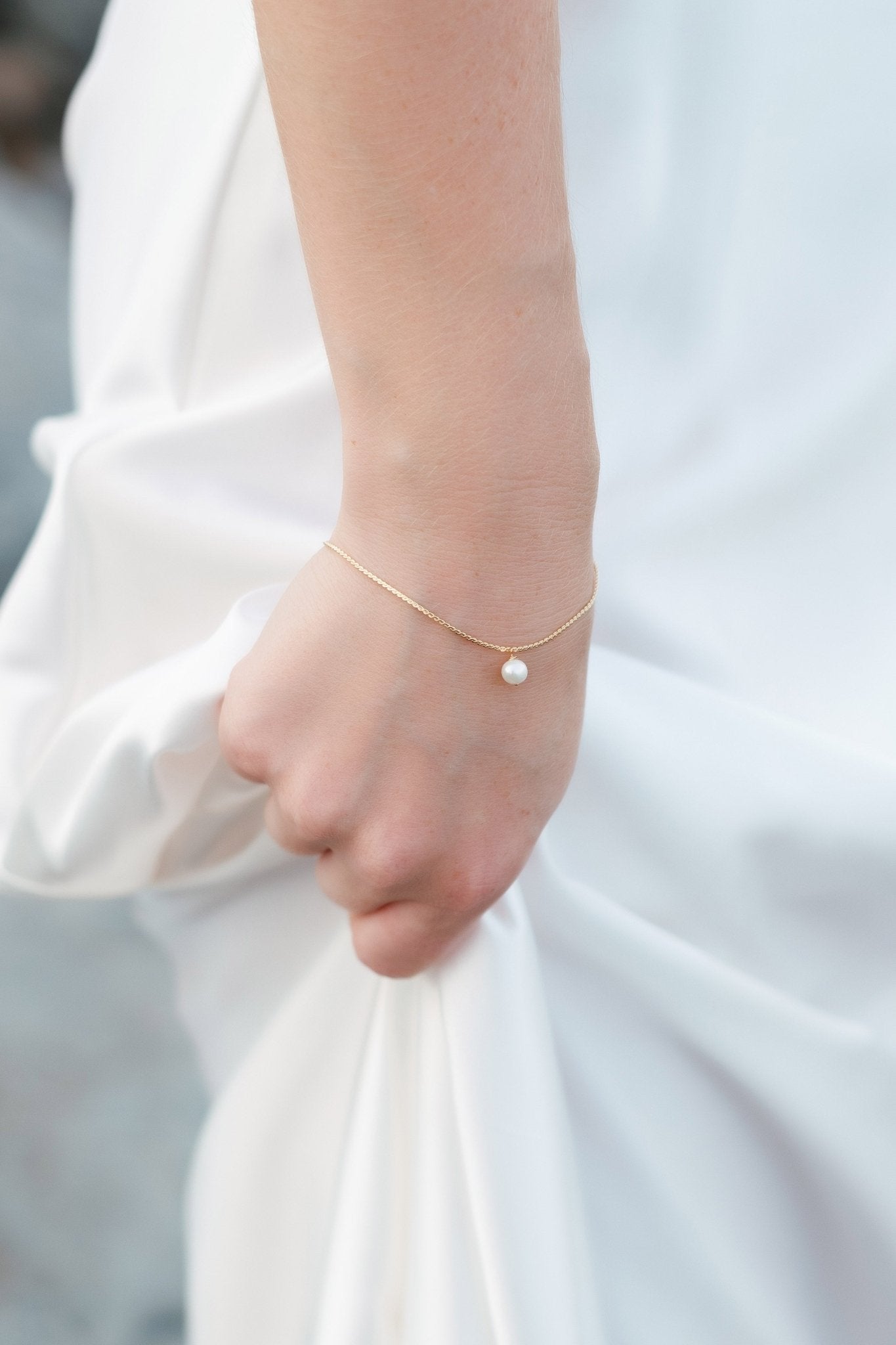 Hand grasps a white satin wedding dress wearing a dainty gold-filled bracelet with floating freshwater pearl. 