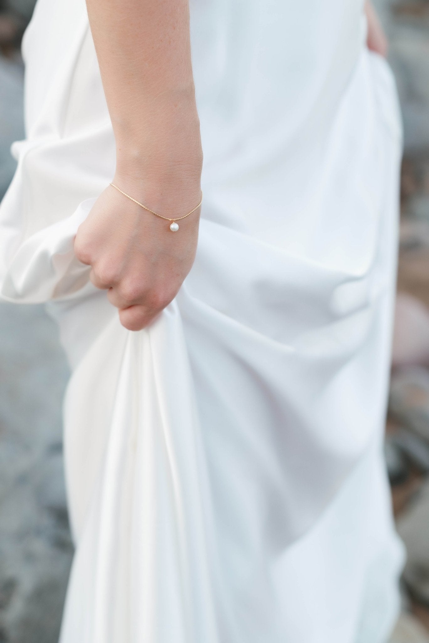 Sleek white wedding gown held up by a hand showcasing a braided gold-filled bracelet with floating freshwater pearl. 