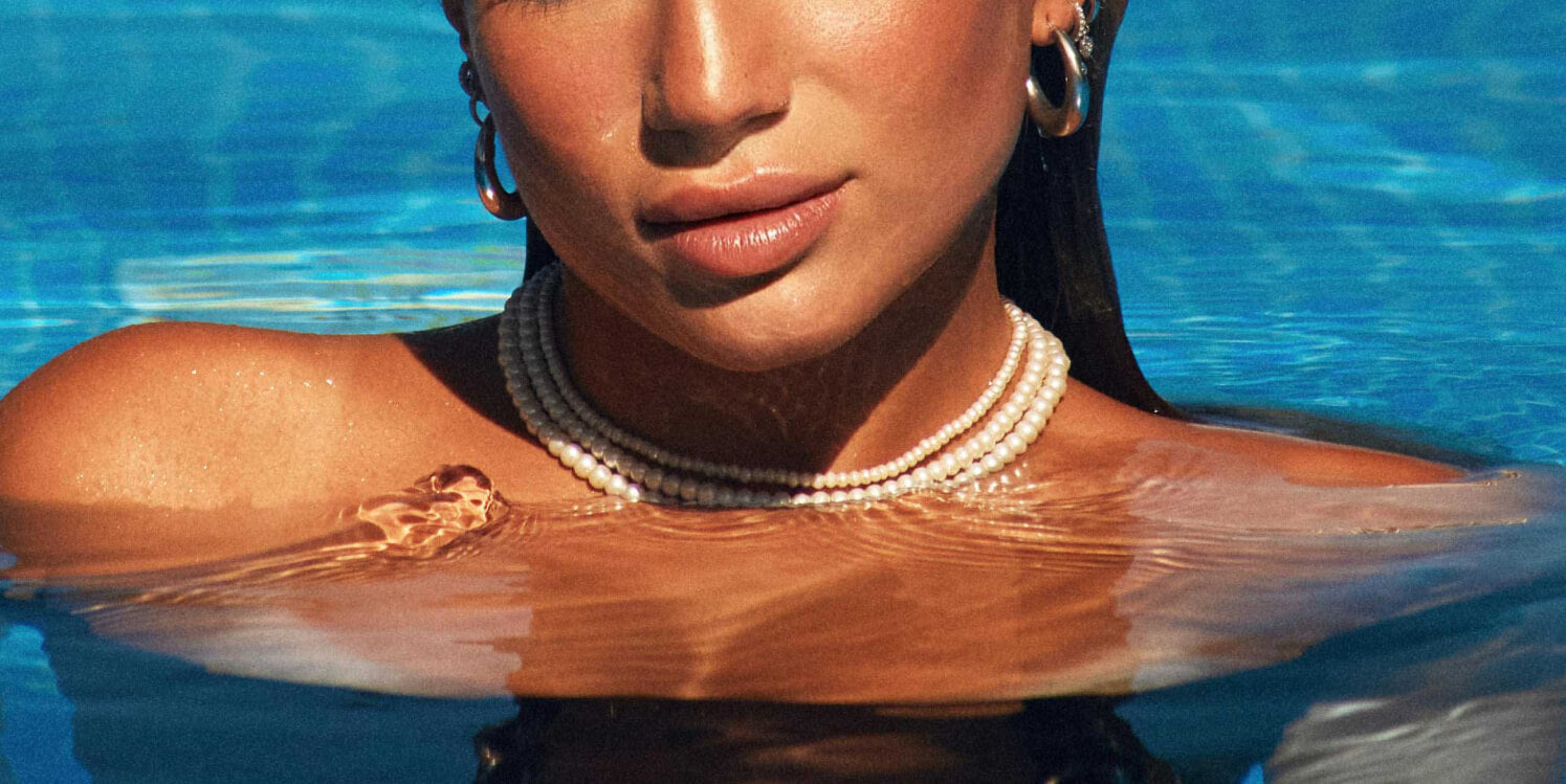 Close up of a woman in a swimming pool wearing several pearl necklaces and gold earrings.