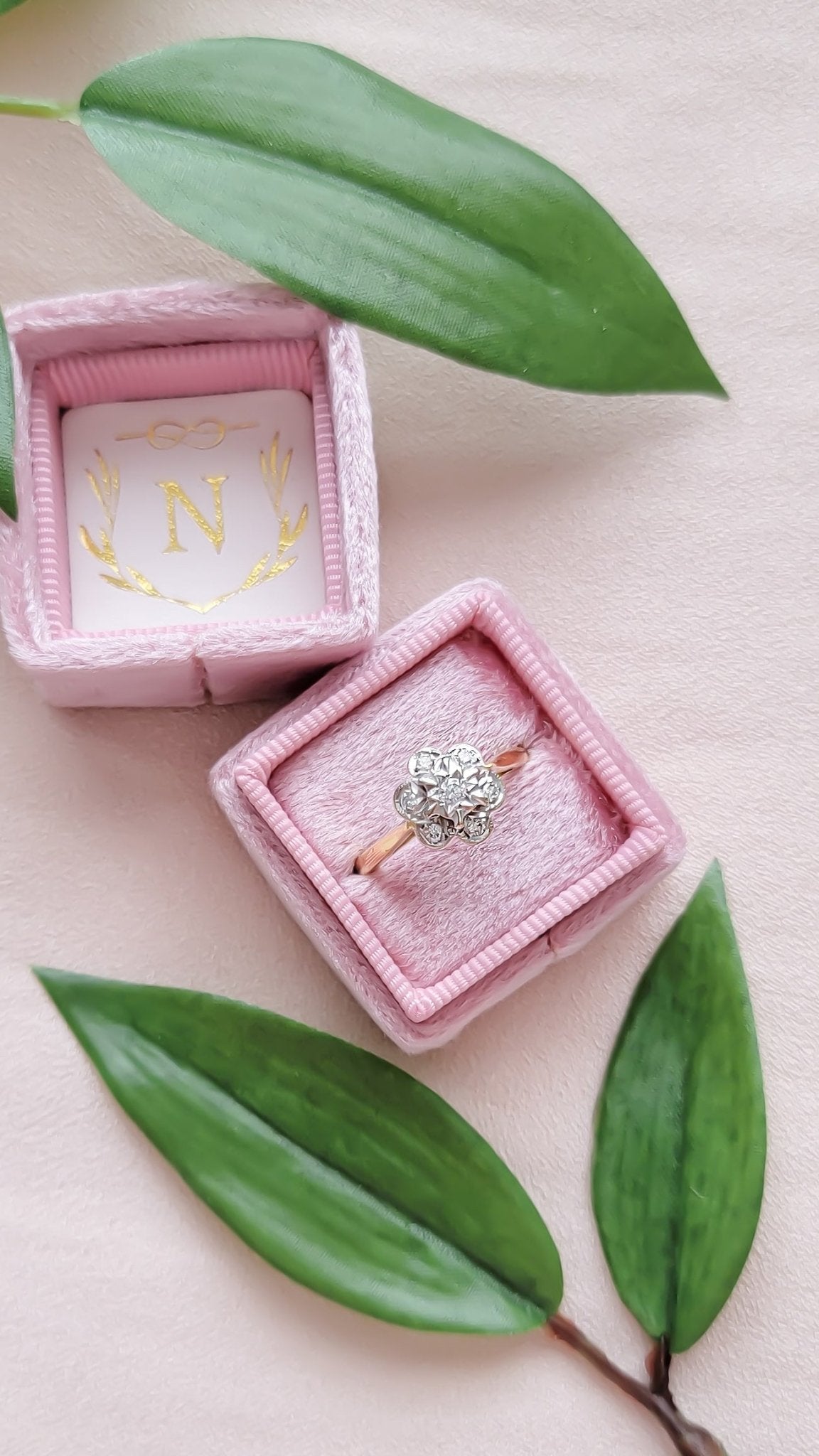 An 18ct Yellow and White Gold Diamond Flower Vintage Engagement Ring sits in a pink square ring box with leaves. 