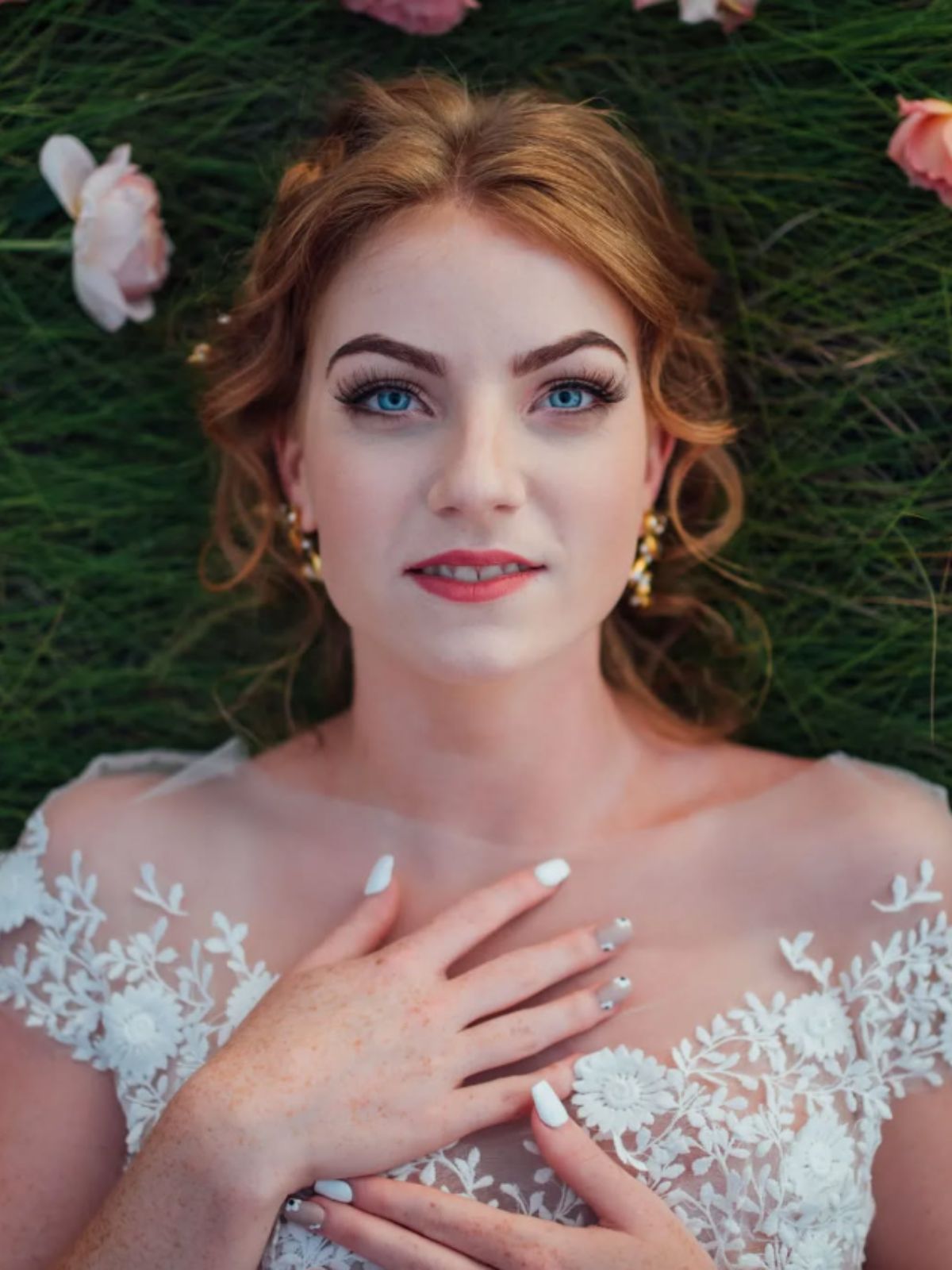 A redheaded bride in a lace wedding dress laying in the grass with flowers. Bridal designer Canada