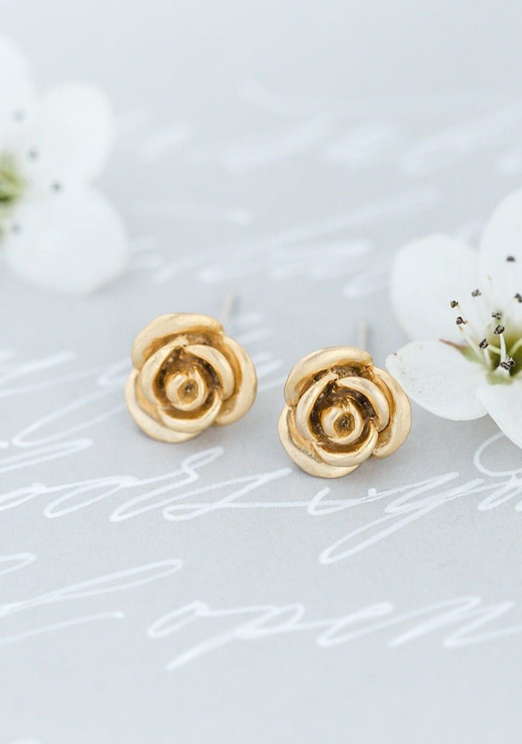 A pair of gilded 14k gold-filled rose studs with sterling silver posts sitting on pale grey surface. Gold-filled jewelry, handmade Canadian jewelry, & affordable jewelry by BIPOC jewelry designer