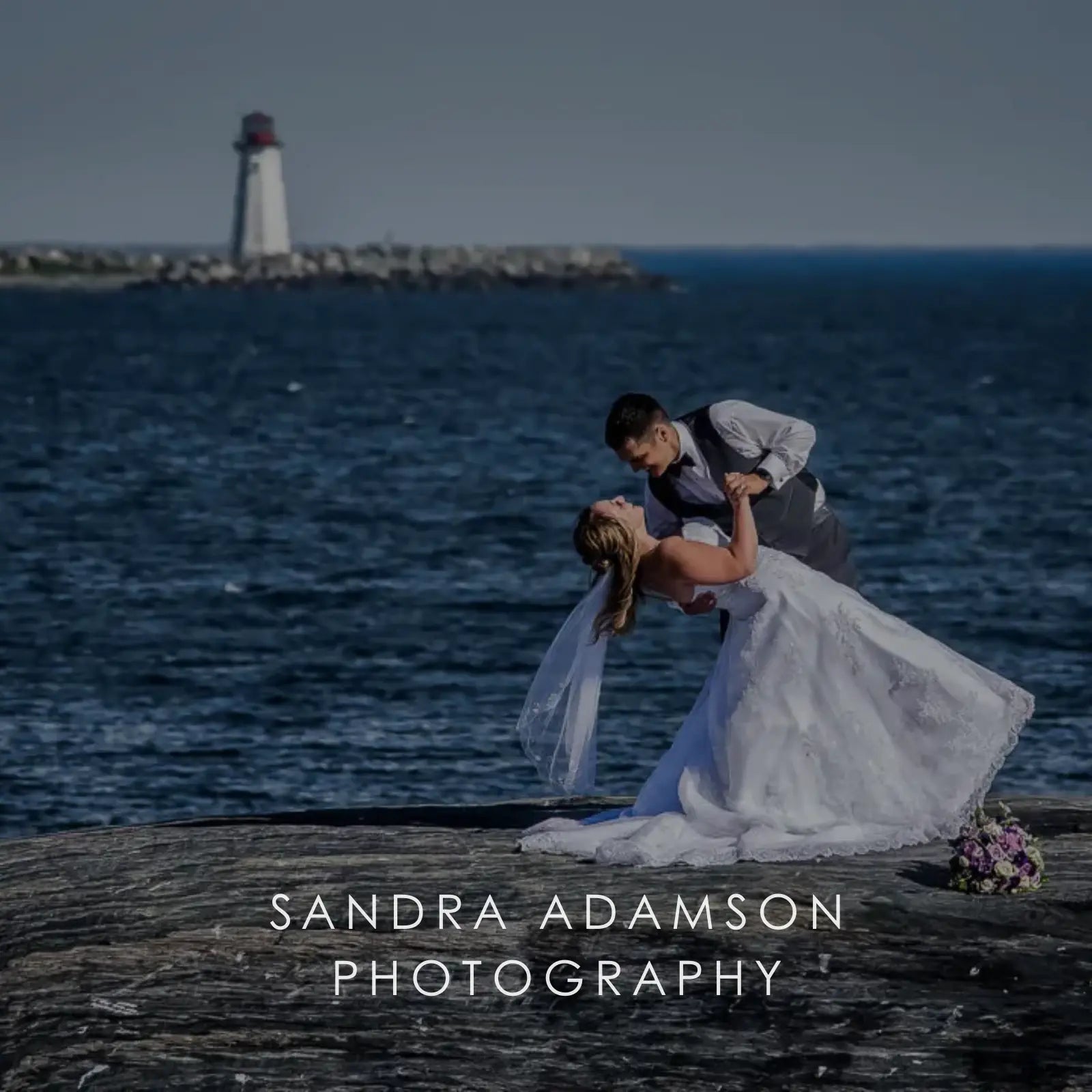 Groom dipping bride by the water with a lighthouse in the background, photo by Sandra Adamson Photography.