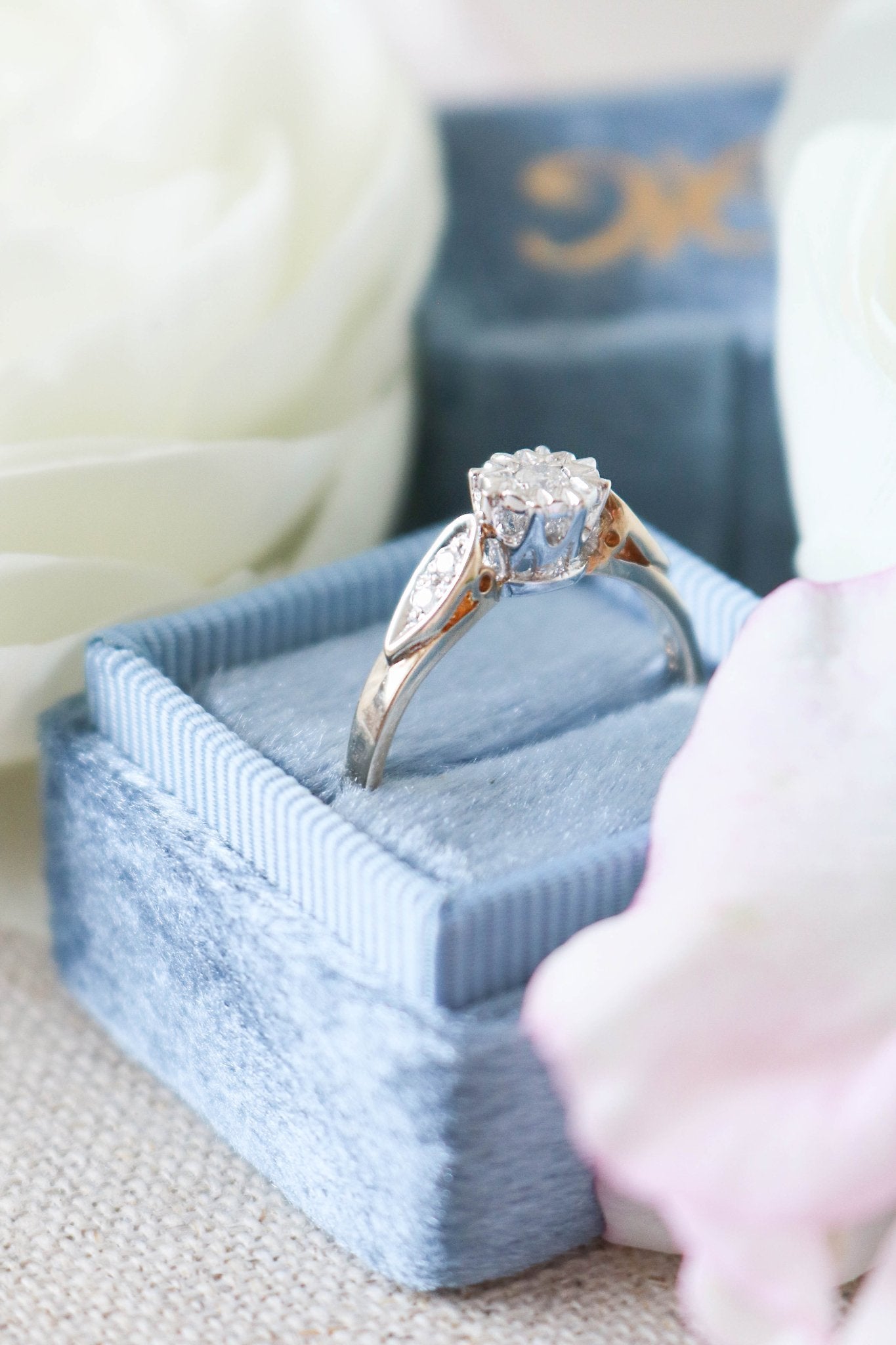 A close up side view of a 9ct White and Yellow Gold Diamond Vintage Engagement Ring in a blue ring box surrounded by white roses. 