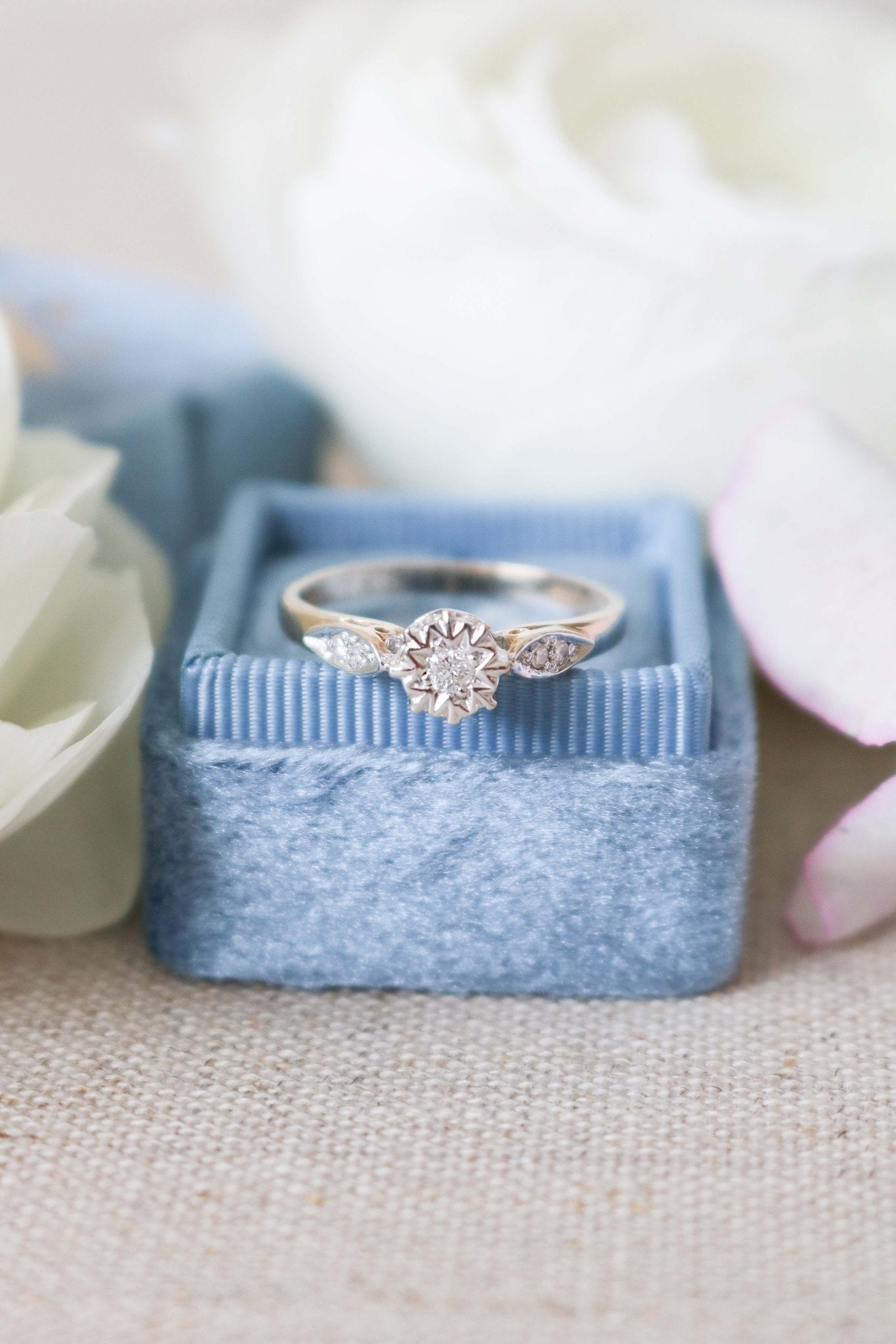 A close up front view of the Serena Diamond Vintage Engagement Ring in 9ct Yellow and White Gold on a blue ring box surrounded by white roses. 