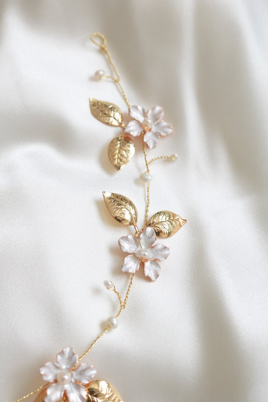 An extreme close up of the gold and enamel flowers with wire-wrapped freshwater pearls against a white satin sheet. 