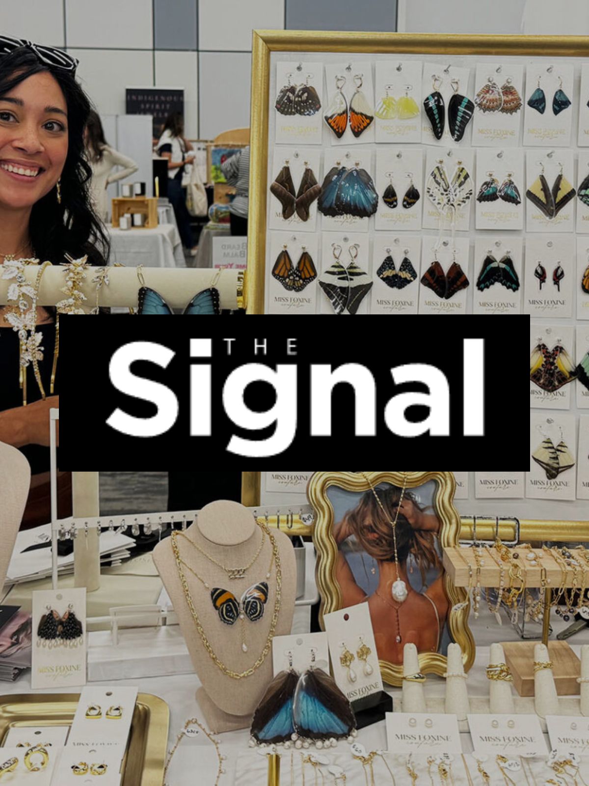 A black and white logo for The Signal article with background of Miss Foxine Couture display featuring butterfly wing earrings, gold-filled bracelets, rings, and freshwater pearl necklaces.
